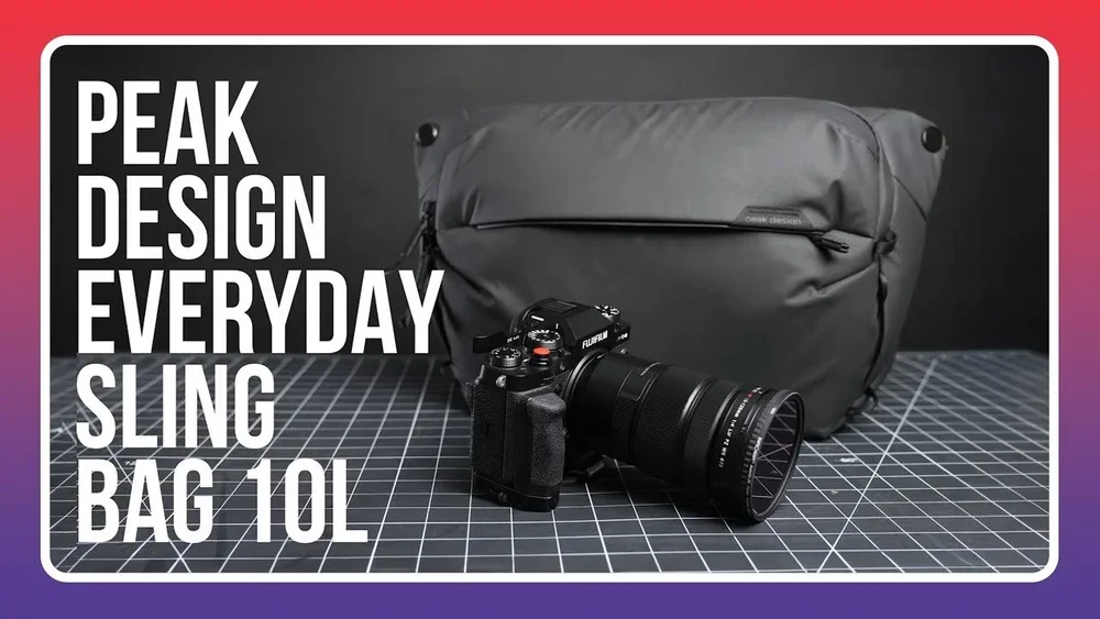A black and gray sling bag with a large logo, placed on a grid-patterned surface, with a professional camera with a large lens in front of it, and a black background. Text on the image reads "Peak Design Everyday Sling Bag 10L."