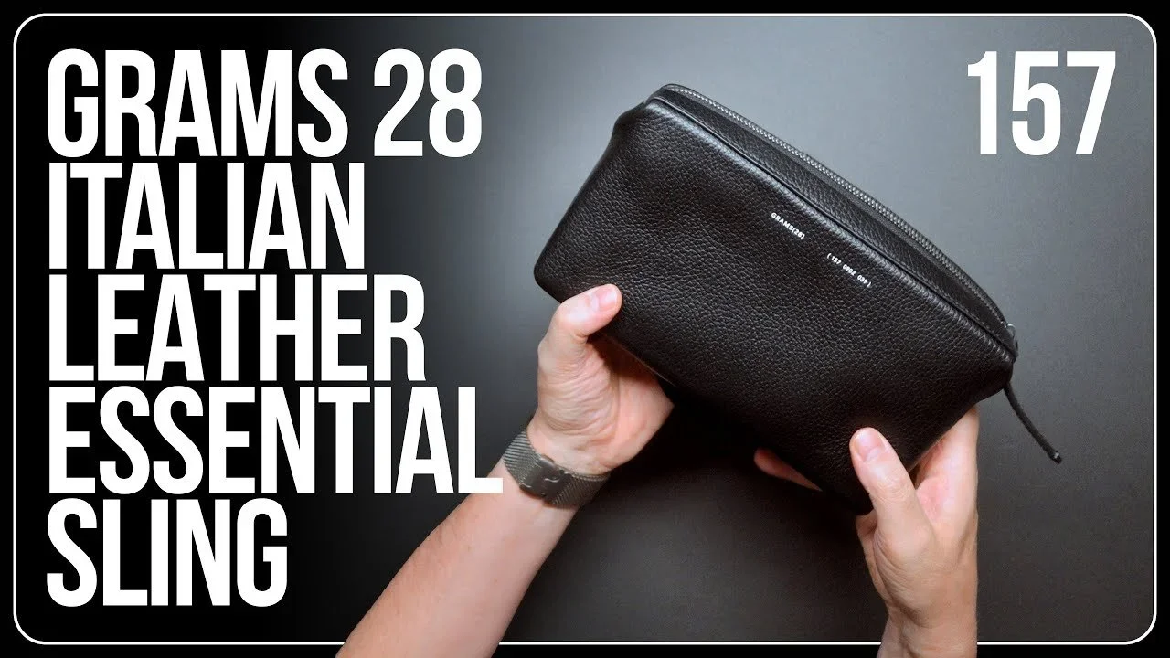 A black leather bag with a zipper held by a person's hands against a gray background.
