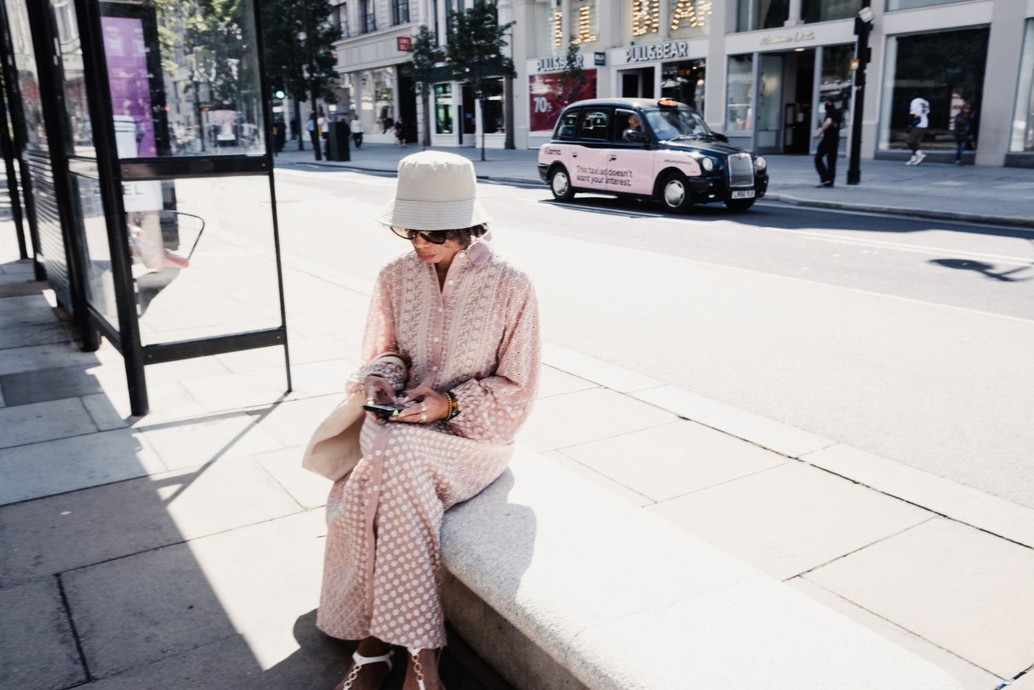 A woman sitting on a stone bench waiting at a bus stop, looking at her phone, in an urban street scene with cars and pedestrians.
