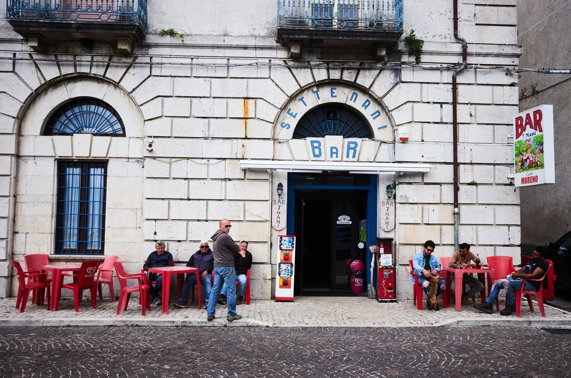 People sitting outside a bar with red tables and chairs in front of a white stone building with blue window frames and balconies. The bar's entrance has signs reading 'SETE NAPOLI' above and 'BAR' below, with additional signs on the walls displaying 