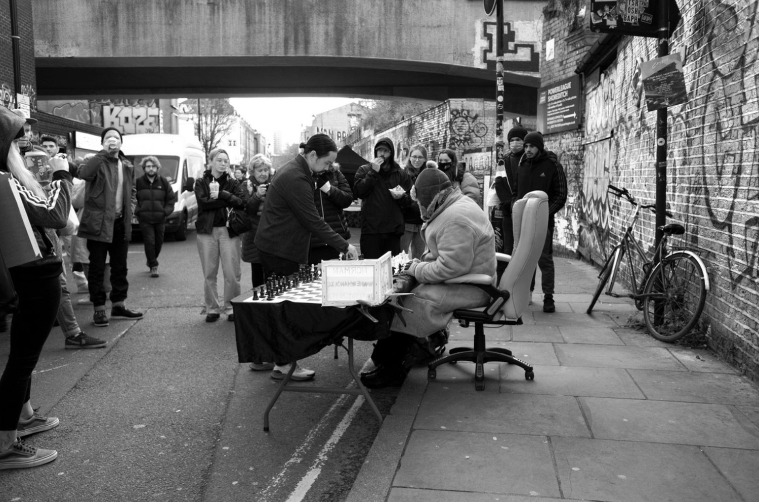 A street scene with a man and woman playing chess outdoors, surrounded by a group of people watching. The scene takes place under a bridge, with graffiti on the walls and a bicycle leaning against the brick wall.