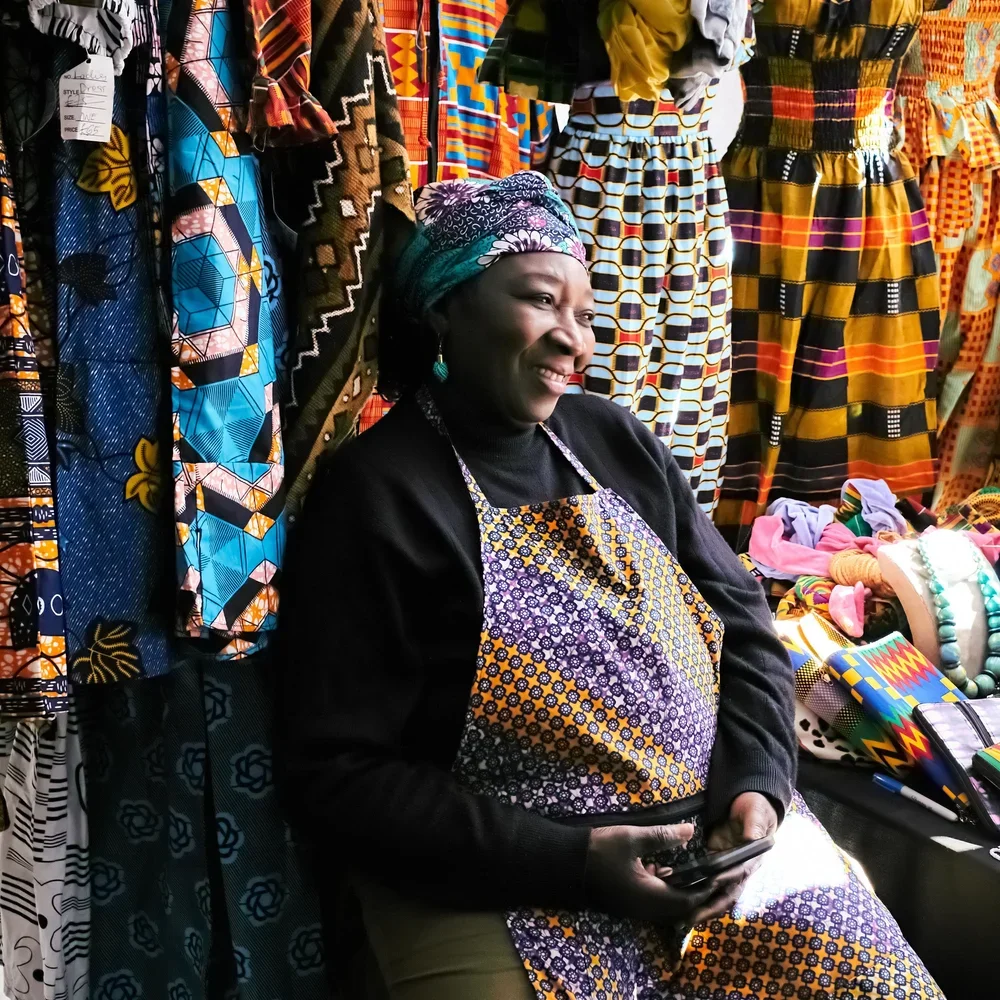 A woman wearing a colorful head wrap and apron sitting at a stall selling vibrant patterned fabrics and accessories.