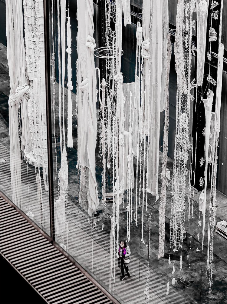 A person standing on a walkway looking up at hanging fabric art installations in a gallery, with various inky textures and patterns on the fabric.