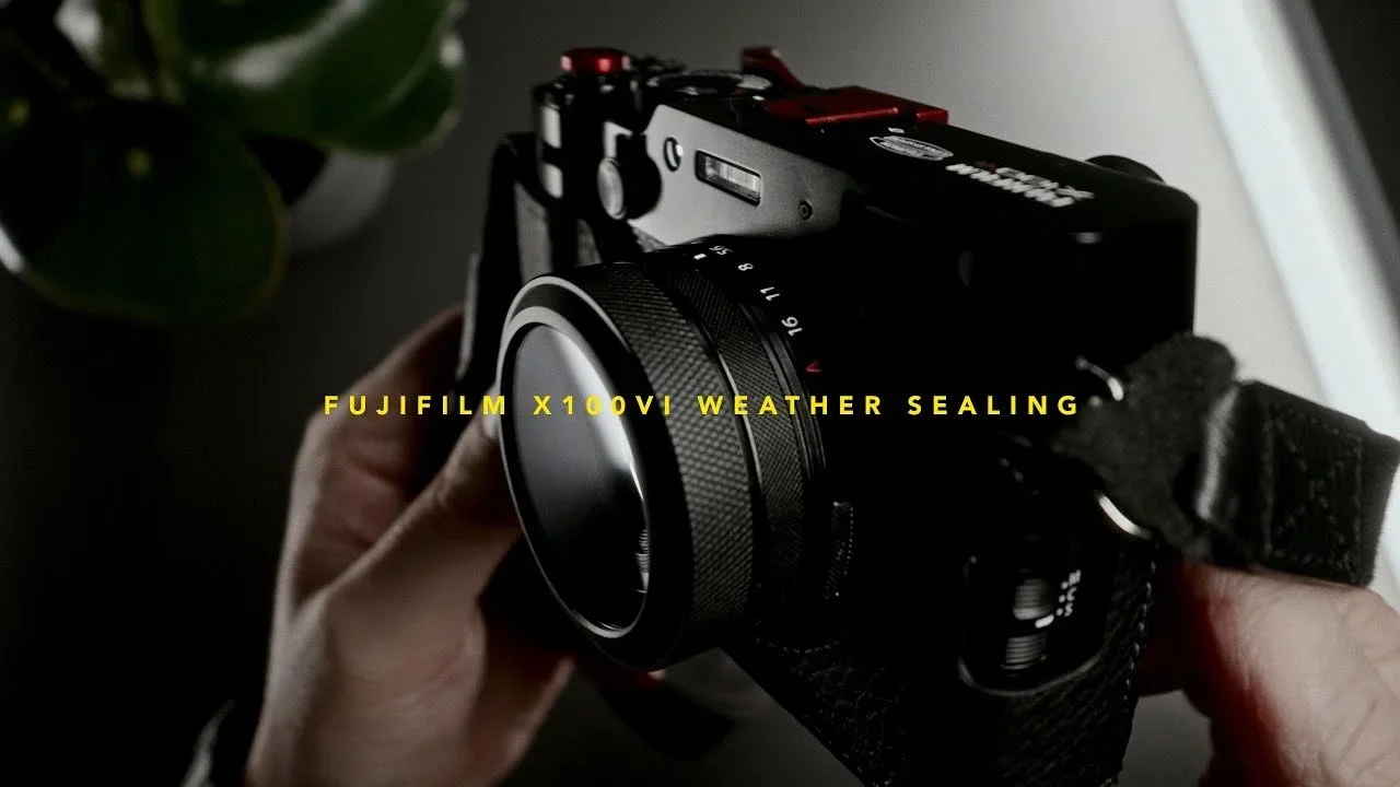 Hand holding a Fujifilm X100V camera with weather sealing, indoors near a window with a plant in the background.