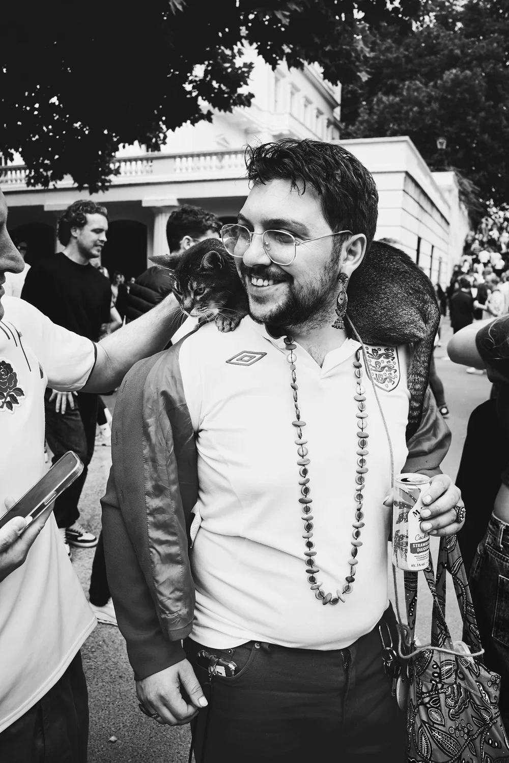 A man smiling with a cat on his shoulder during a outdoor gathering.