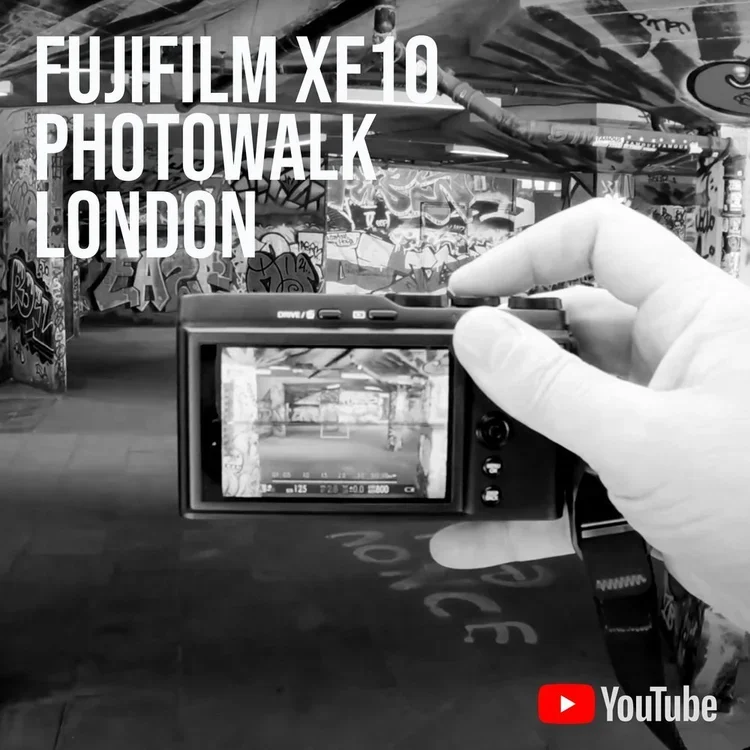 A person holding a digital camera in front of graffiti-covered walls at a London photo walk event for Fujifilm XE10, with a YouTube logo in the bottom right corner.