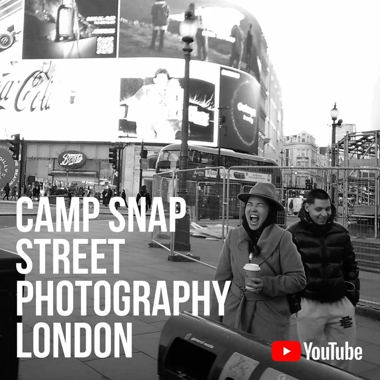 Black and white photo of a busy street scene in London with two smiling people, one holding a coffee cup, and urban advertisements on buildings, with the text 'Camp Snap Street Photography London' and a YouTube logo.