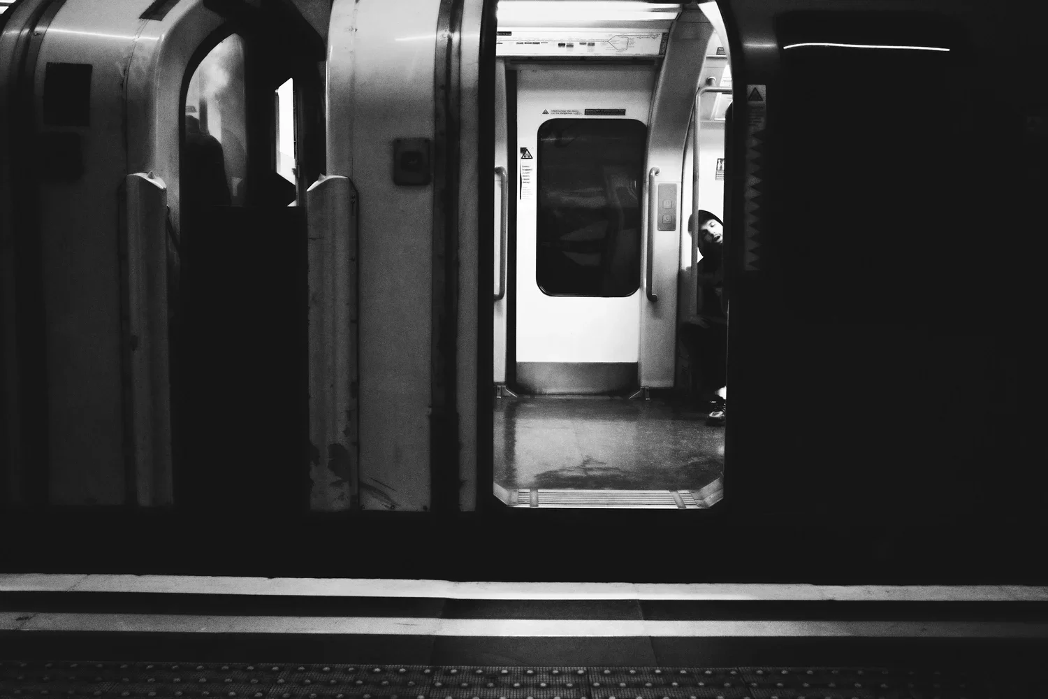 Black and white photo of an empty subway train station platform with a train door open. Inside the train, a person with a beard is seated near the door, looking towards the camera.