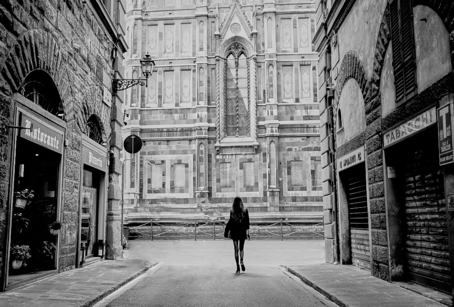 A woman walking down a narrow European street with old stone buildings, with a large historic cathedral or church in the background.