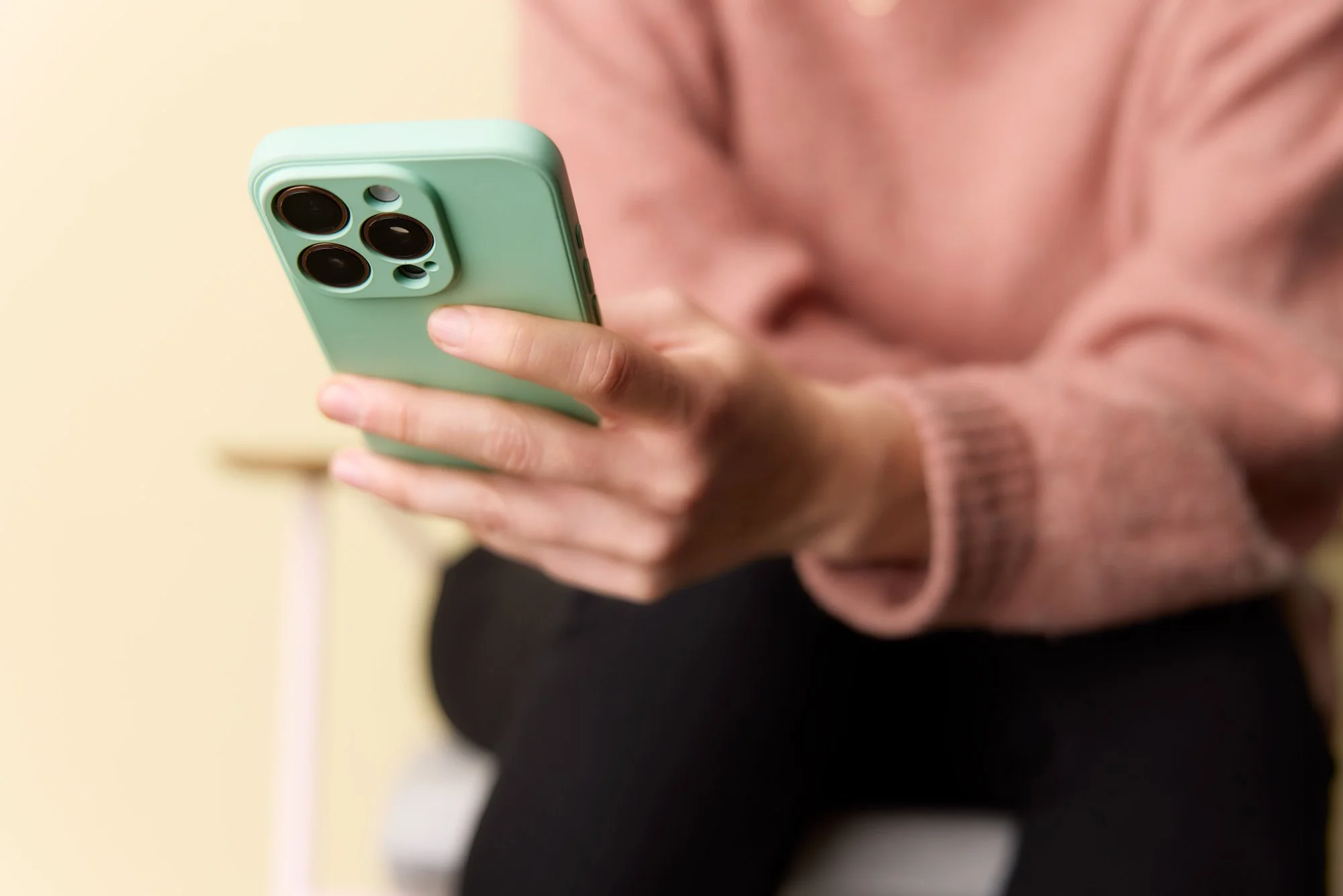 Person hält ein mintgrünes Smartphone mit mehreren Kameralinsen, trägt eine pinke Strickjacke und schwarze Hosen, im Hintergrund beige Wand.