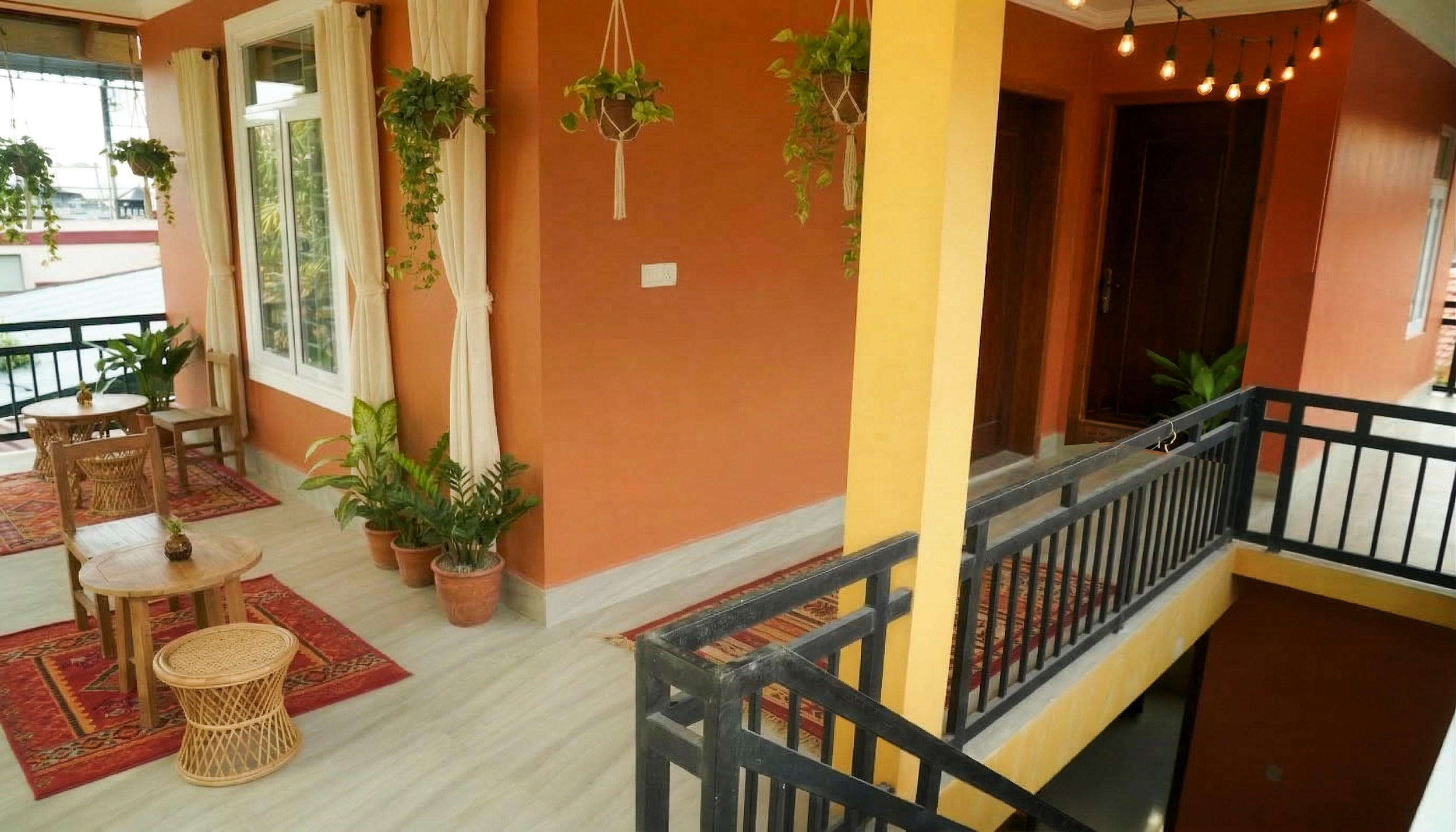 Interior of a brightly lit room with orange and yellow walls, decorated with hanging potted plants and potted plants on the floor. There are wooden tables and chairs with red and orange patterned rugs, and large windows draped with white curtains. A staircase with a black railing leads down from the right side.