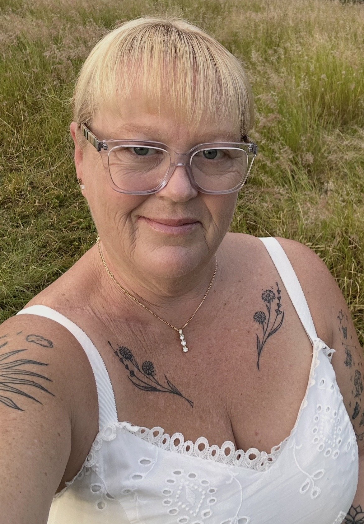 A woman with short blonde hair wearing large glasses, a white eyelet dress, a gold necklace with a pendant, and visible tattoos on her shoulders and arms, standing outdoors on grass.