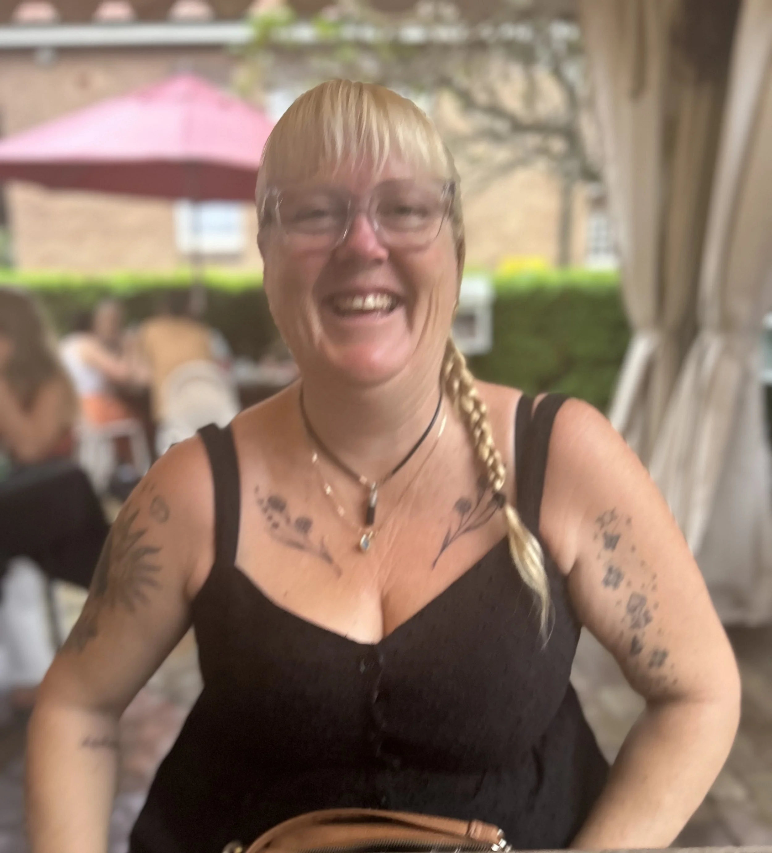A smiling woman with blond hair in a braid, wearing glasses, a black sleeveless top, and tattoos on her arms and chest, sitting outdoors at a restaurant with a pink umbrella and other people in the background.