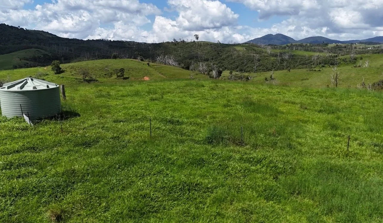 Green rolling hills with scattered trees under a partly cloudy sky, and a large metal water tank in the foreground on the left.