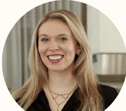 A smiling young woman with long blonde hair, wearing a black top and layered necklaces, standing in a modern indoor setting.