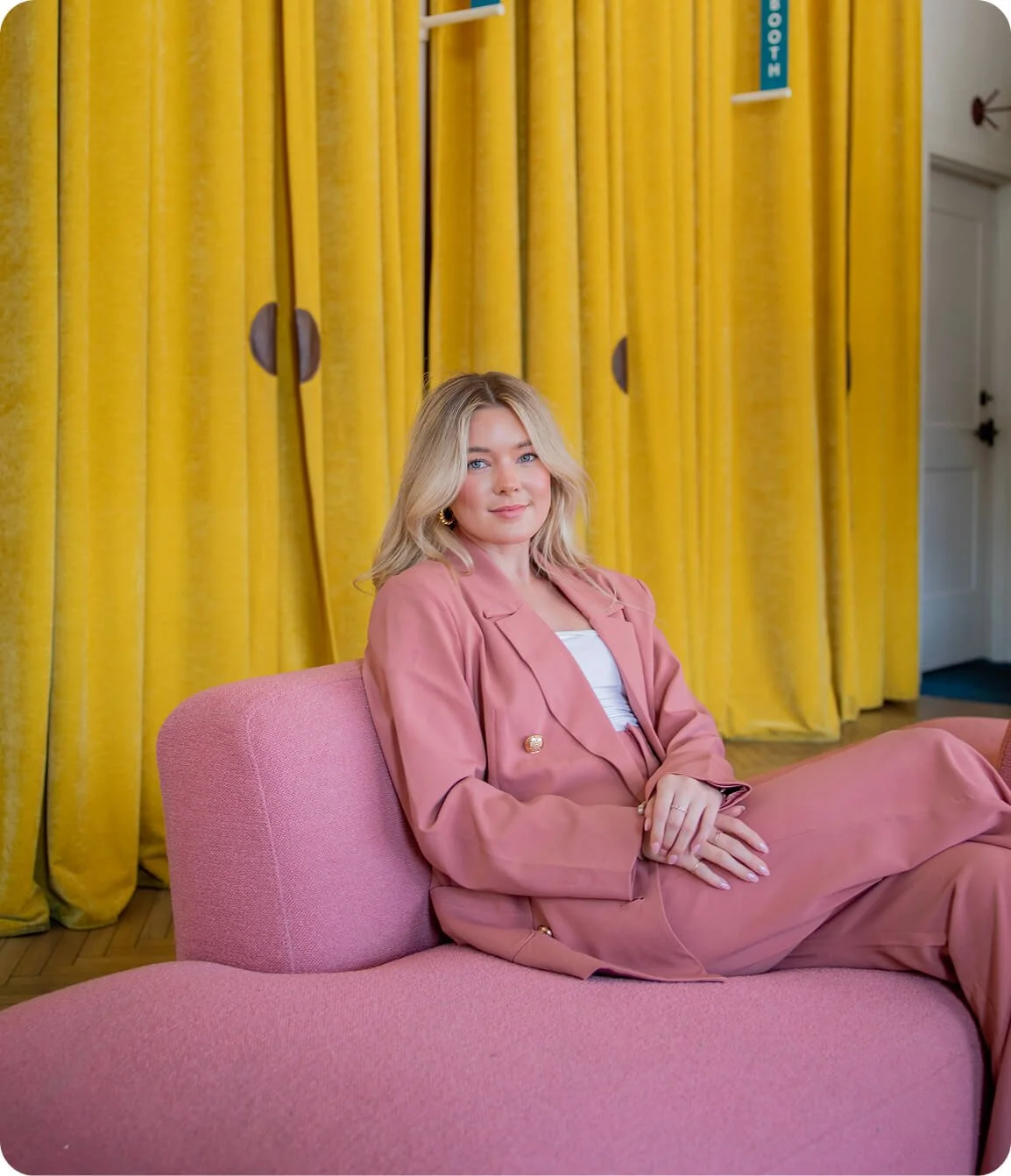 A woman with blonde hair and blue eyes sitting on a pink sofa in front of yellow curtains.