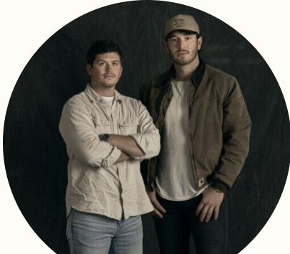 Two young men standing side by side, one with arms crossed and the other with hands in pockets, against a dark background.