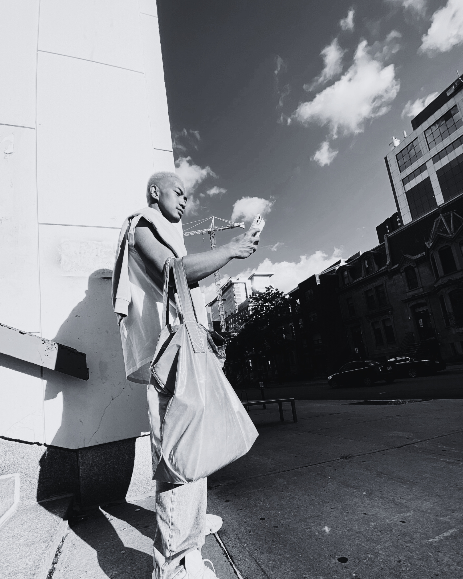 A person standing on a sidewalk against a white wall, looking at a phone, with a cityscape of buildings and a crane in the background, under a partly cloudy sky.