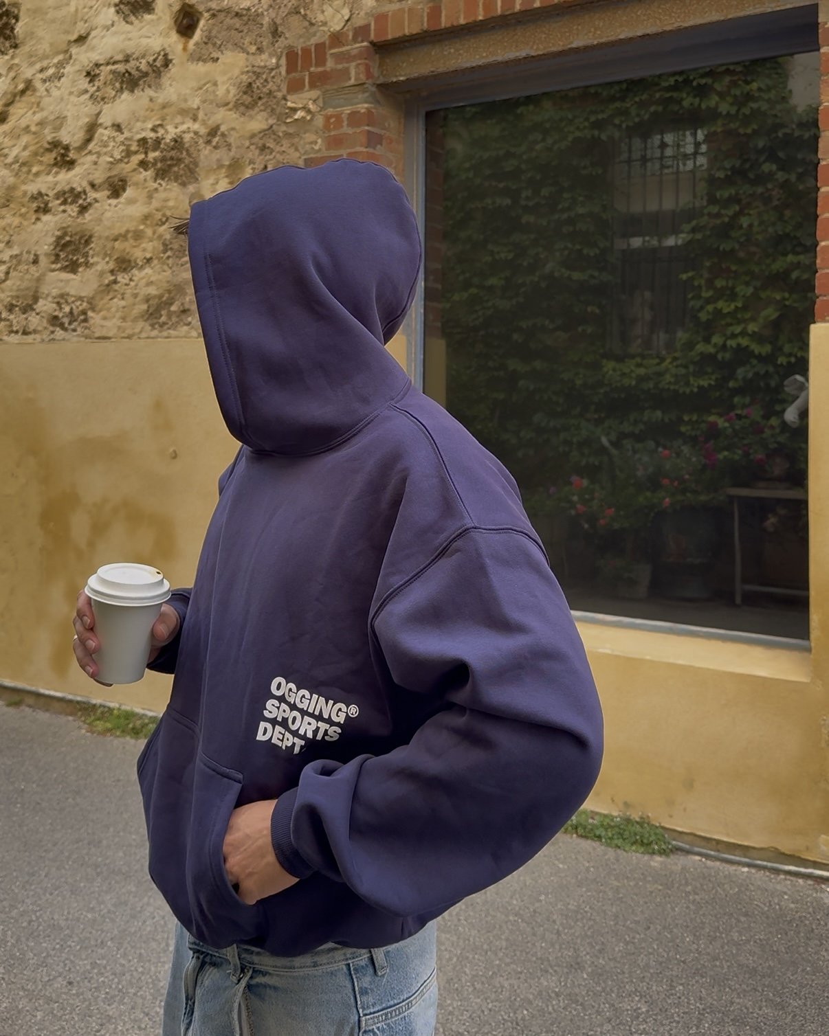 Person wearing a navy hoodie with 'OGGING SPORTS DEPT' printed on it, holding a coffee cup, standing outdoors near a brick and stone wall with a window and greenery in the background.