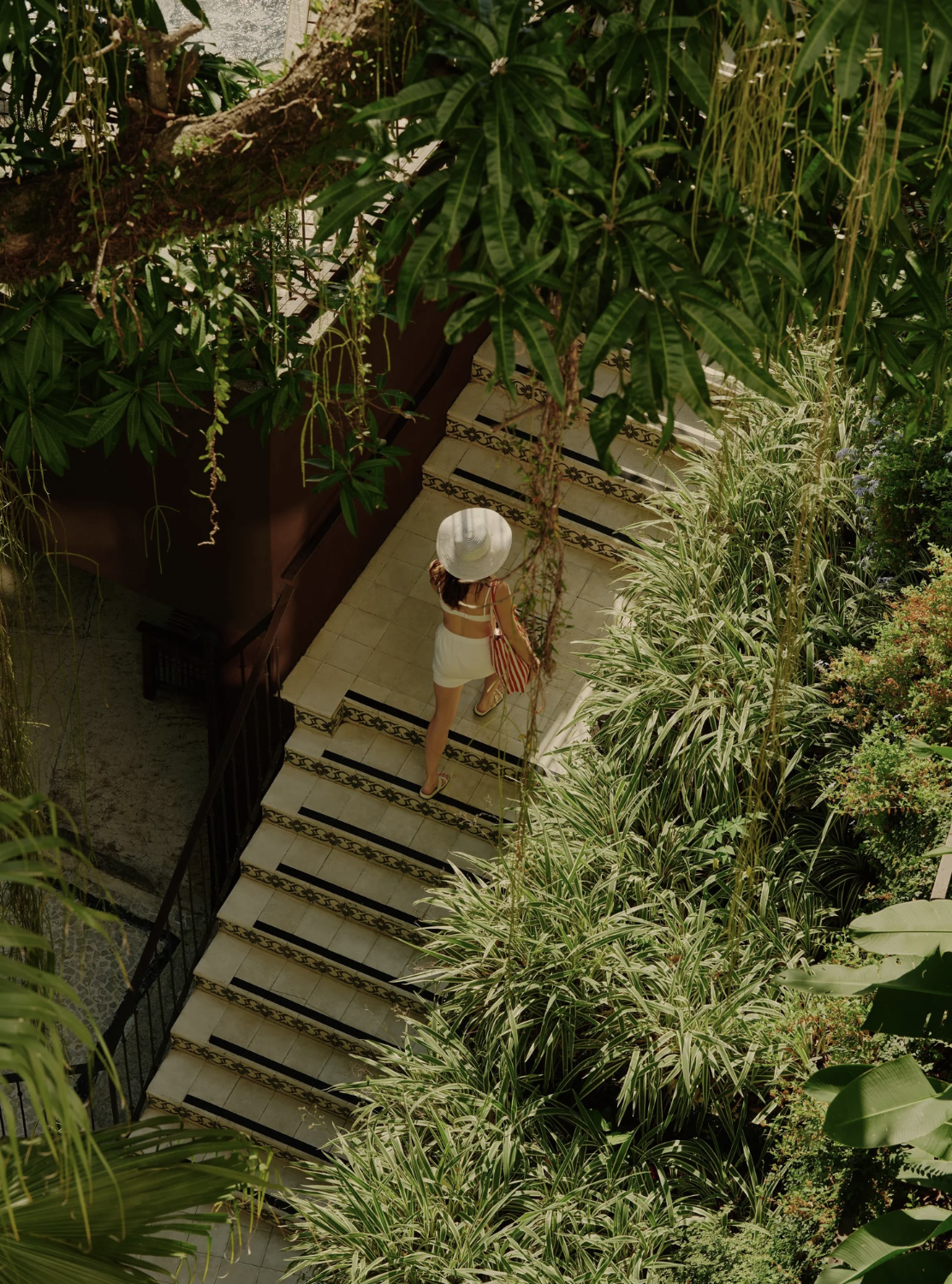 A woman in a white hat and white shorts walking up a staircase through a lush green garden with tropical plants.