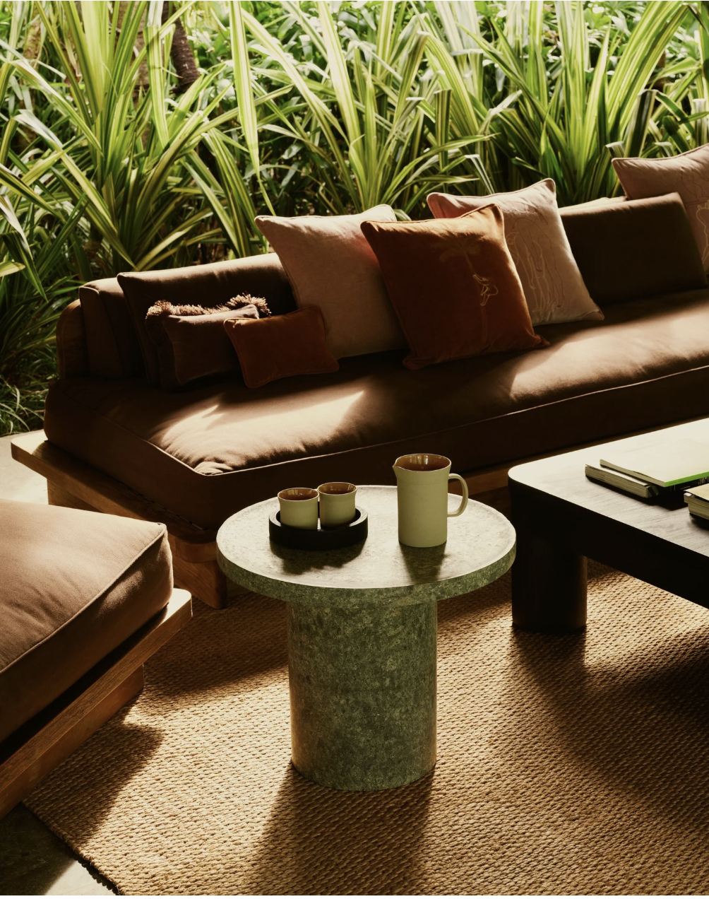 Living room with a brown sofa and several pillows, a circular stone coffee table with a coffee mug and cups, a larger dark coffee table with books, a beige area rug, and a backdrop of tall green tropical plants.