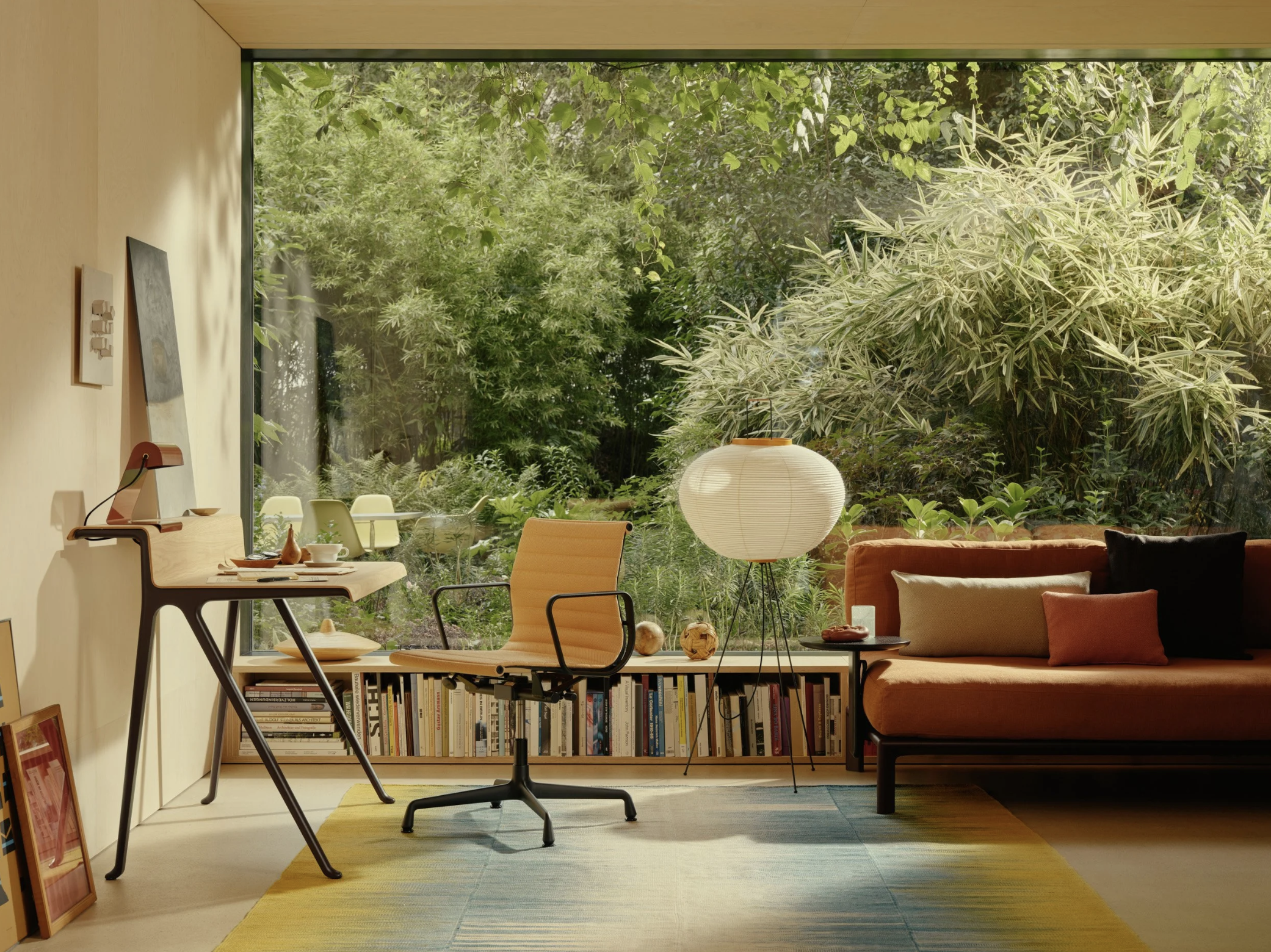 Living room with large window overlooking a lush green garden, featuring a beige sofa with colorful pillows, a white floor lamp, a wooden desk with a chair, and a bookshelf under the window.