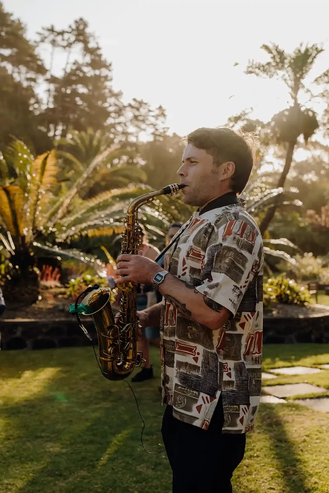 A man playing a saxophone outdoors during sunset, wearing a patterned short-sleeve shirt, with trees and sunlight in the background.
