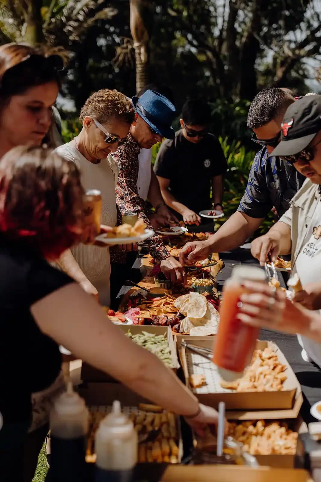 People gathered around a long outdoor buffet table with various food items during daytime.