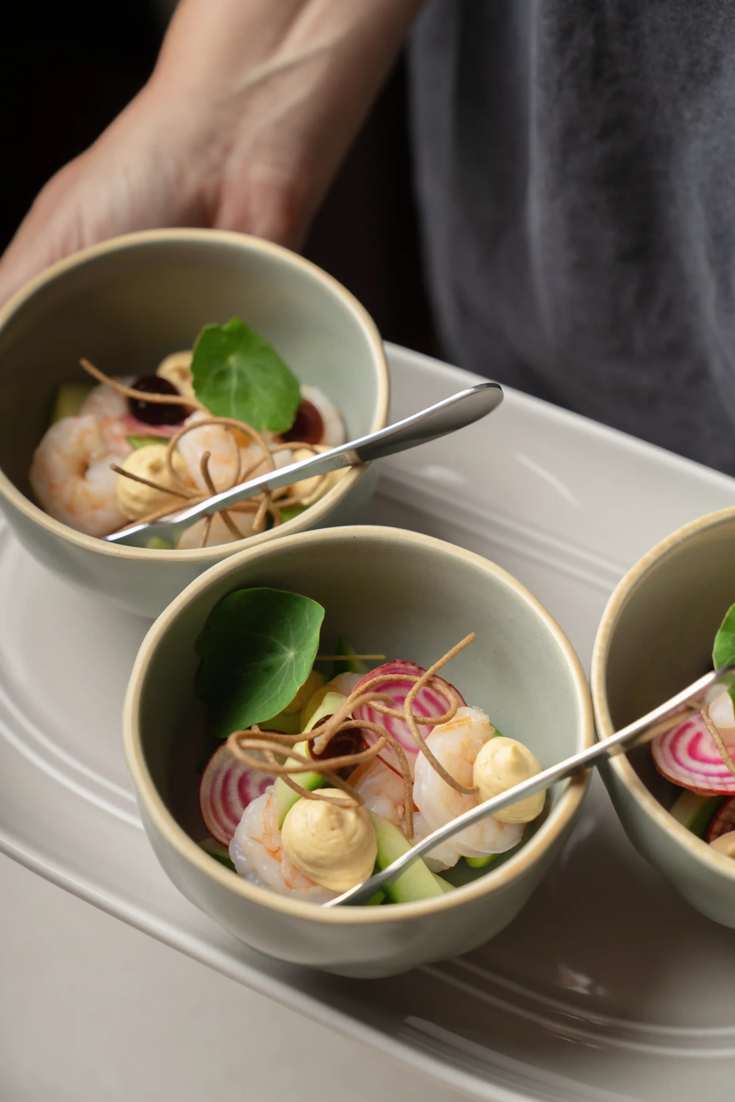 Small cups filled with a shrimp salad garnished with cucumber slices, radish slices, microgreens, and a decorative element, placed on a white tray.
