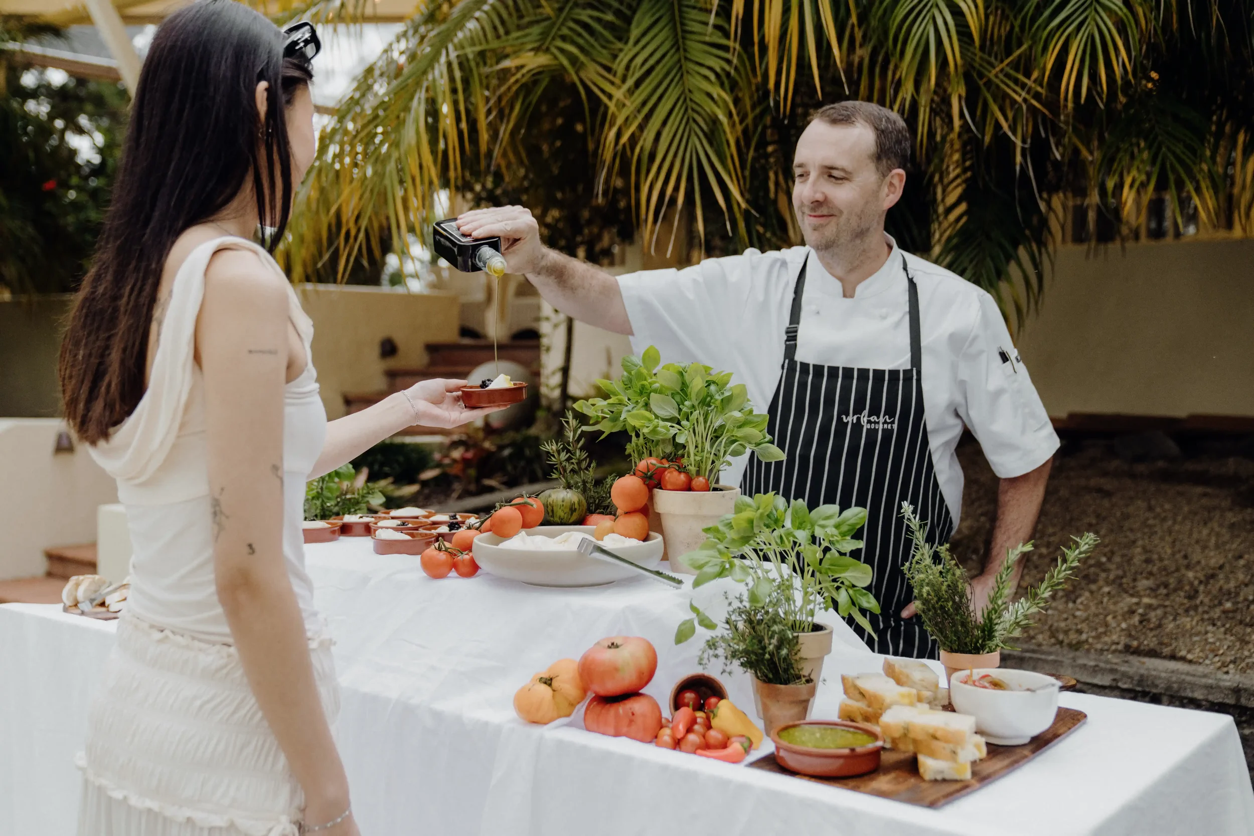 A chef serving a woman at an outdoor food display table with fresh vegetables and prepared dishes, surrounded by trees.