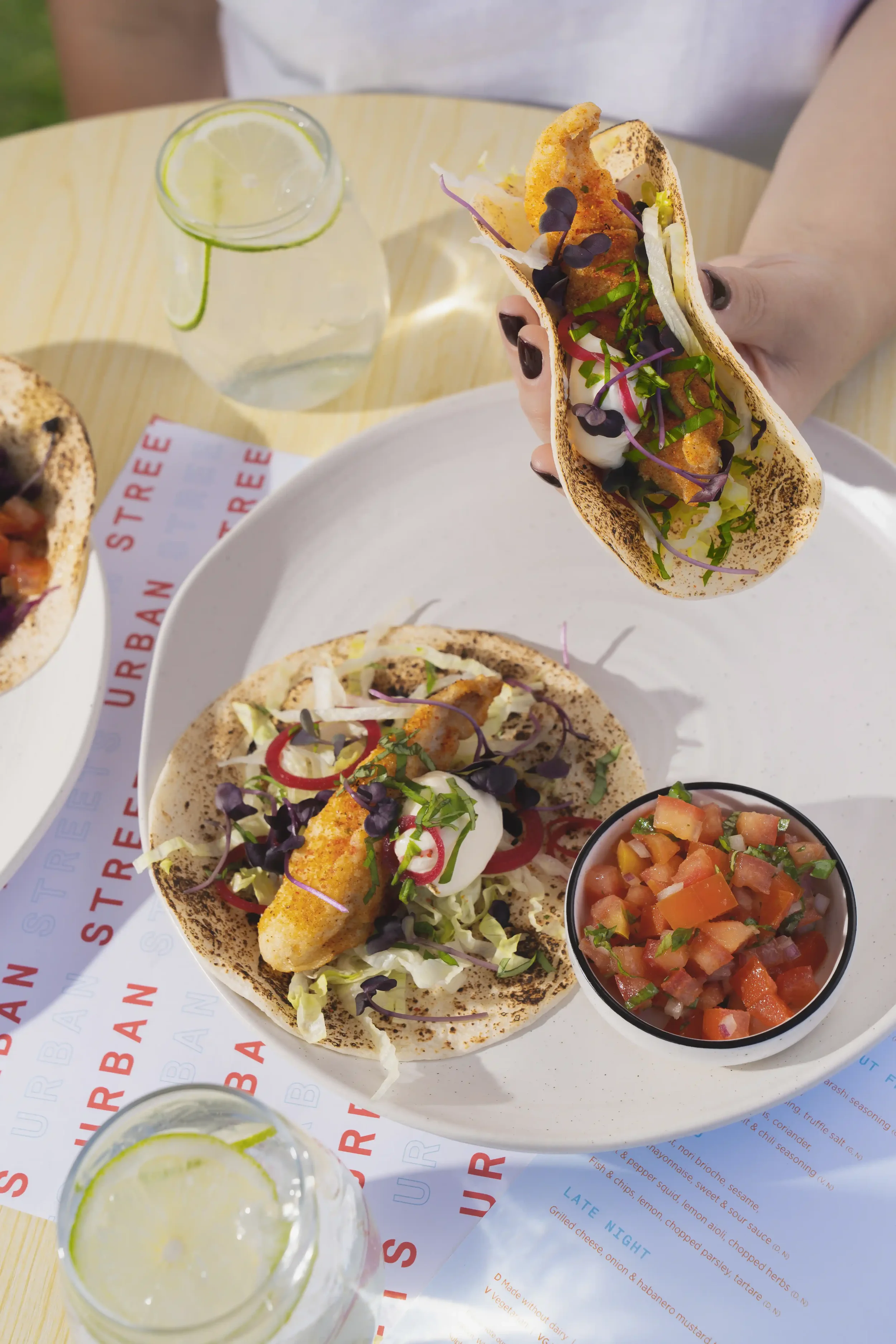 A plate of three tacos filled with fried fish, shredded lettuce, microgreens, and sauce, accompanied by a small bowl of pico de gallo salsa and a glass of lemon-infused water on a yellow table with a menu underneath.