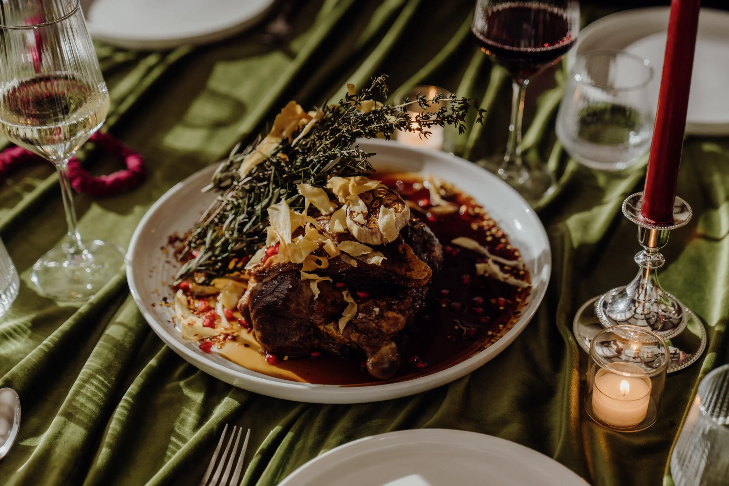 A festive table setting with a large white plate of glazed meat topped with herbs, garlic, and sliced almonds, surrounded by wine glasses, candles, and decorative elements on a green velvet tablecloth.