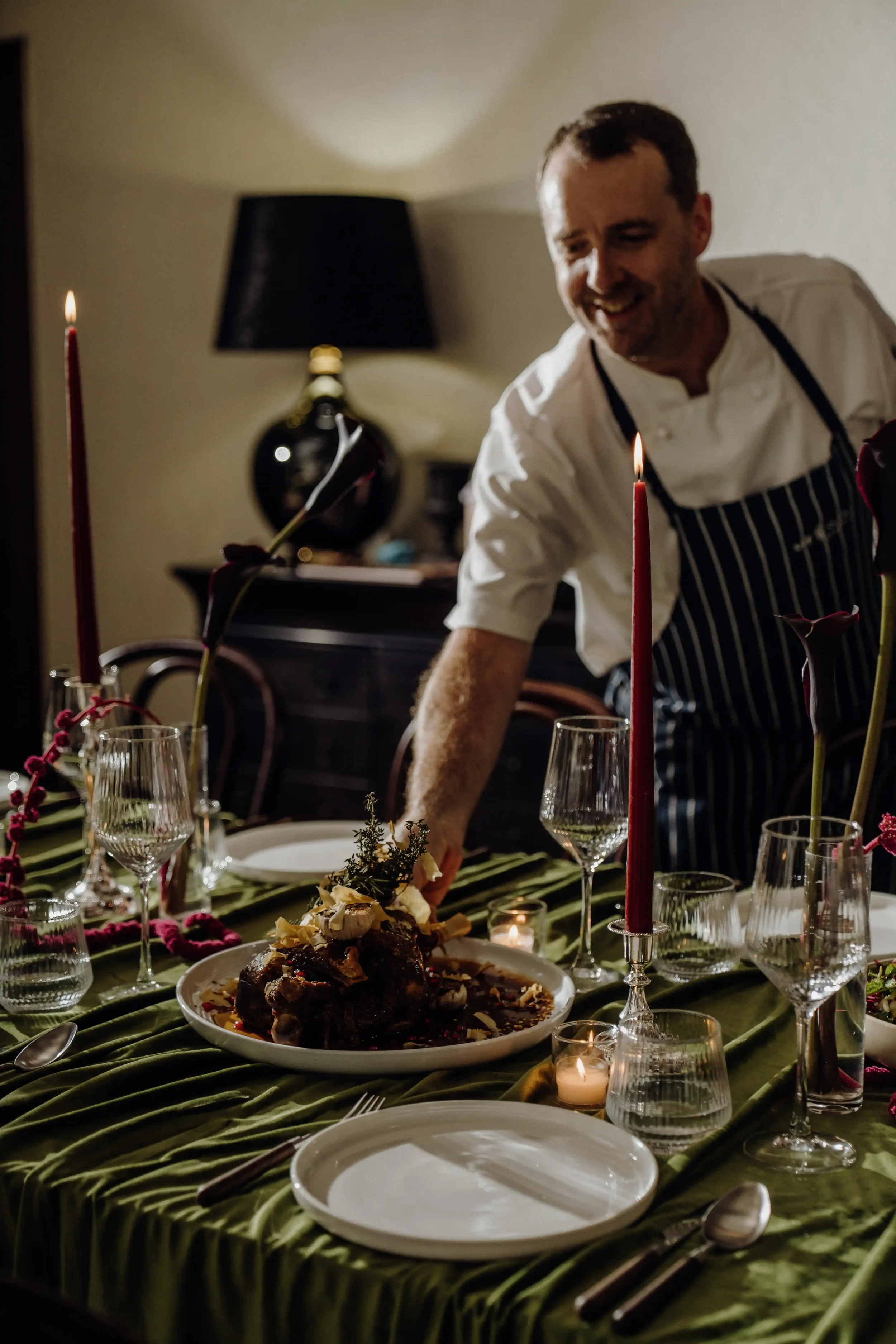 A man in a chef's uniform and striped apron is smiling as he prepares food on a decorated dining table with candles, wine glasses, and floral decorations in a warmly lit room.