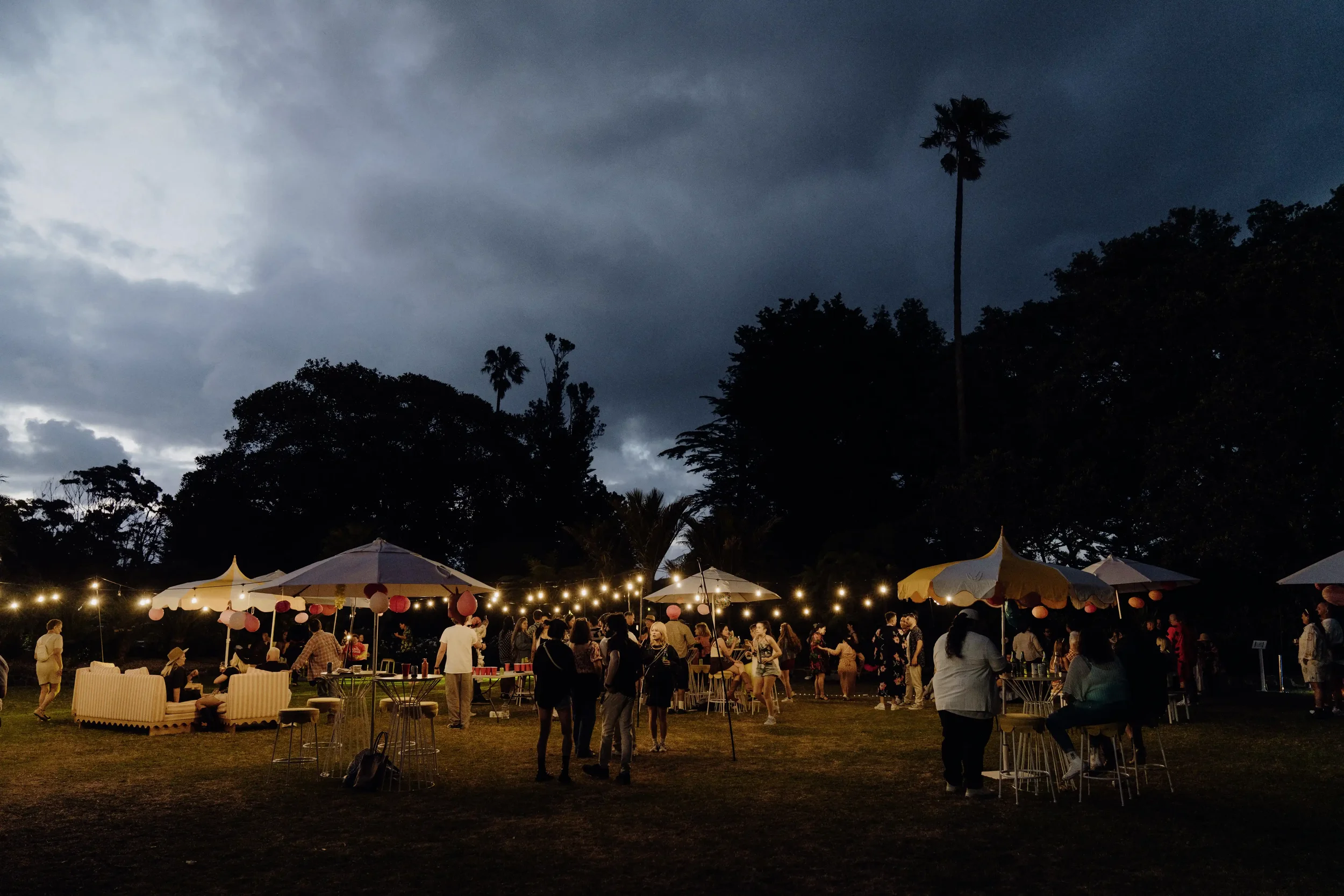 People gathered outdoors during an evening event with string lights, umbrellas, and trees beneath a cloudy sky.
