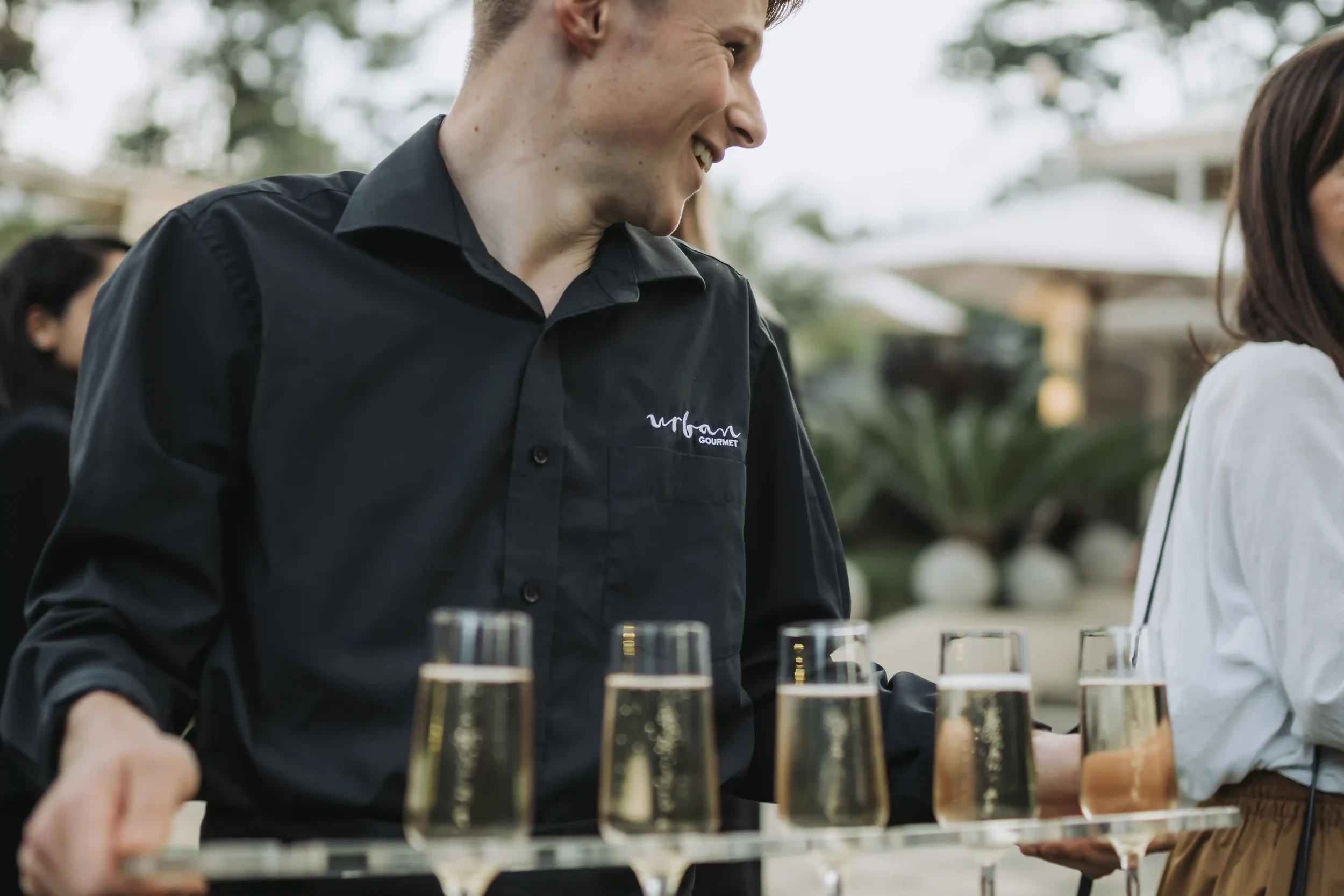 A man in a black shirt smiling at an outdoor event with glasses of beer on a table in front of him.