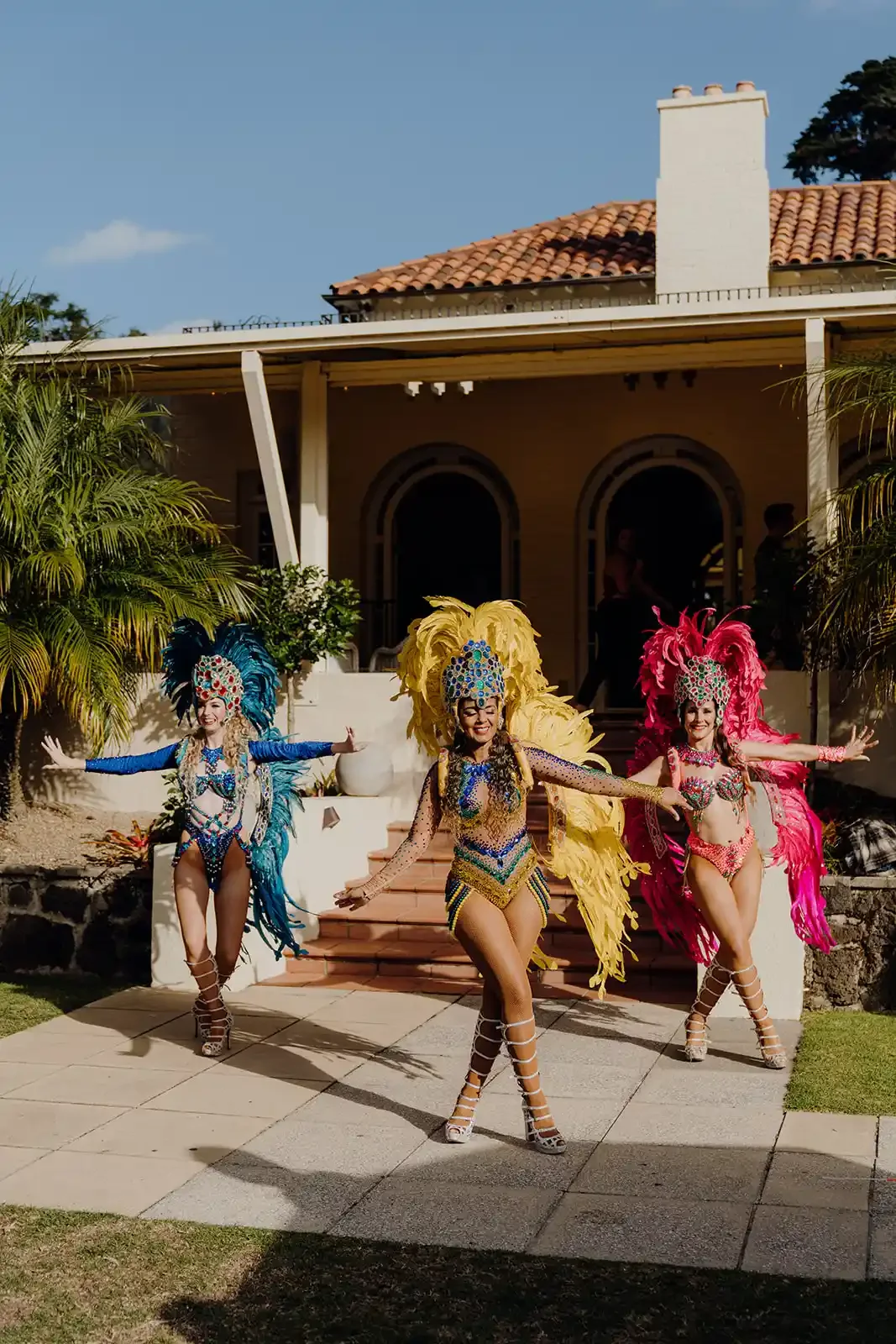 Three women in colorful carnival costumes performing a dance outside a house with a red-tiled roof, palm trees, and a blue sky.