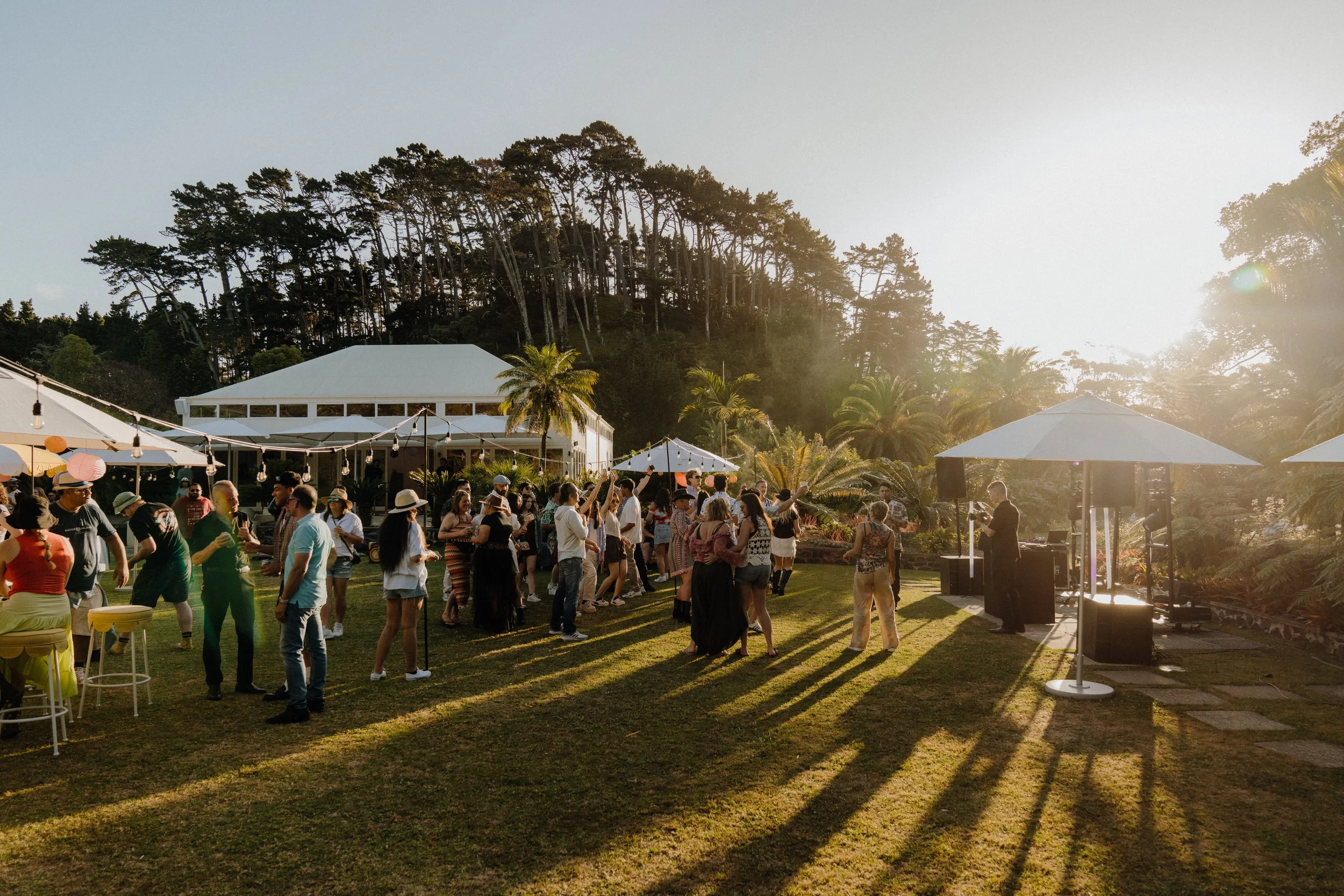 People dancing and socializing outdoors at a yard party or gathering during sunset with tents and string lights, surrounded by trees and hills.
