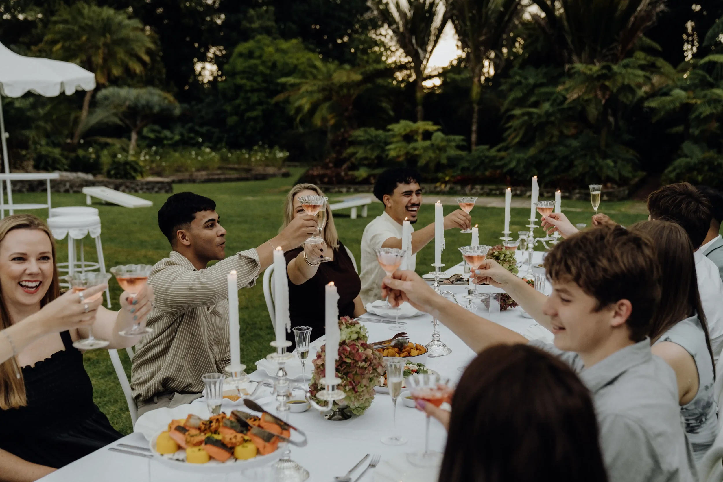 People sitting at a long table outdoors, raising glasses for a toast during an evening gathering, with candles and food on the table, surrounded by lush greenery.