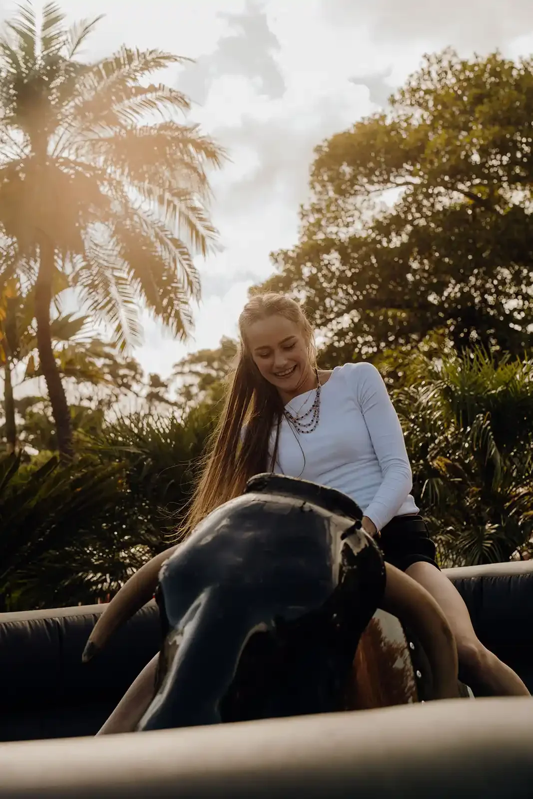 Young woman smiling while riding a mechanical bull outdoors with palm trees and cloudy sky in the background.