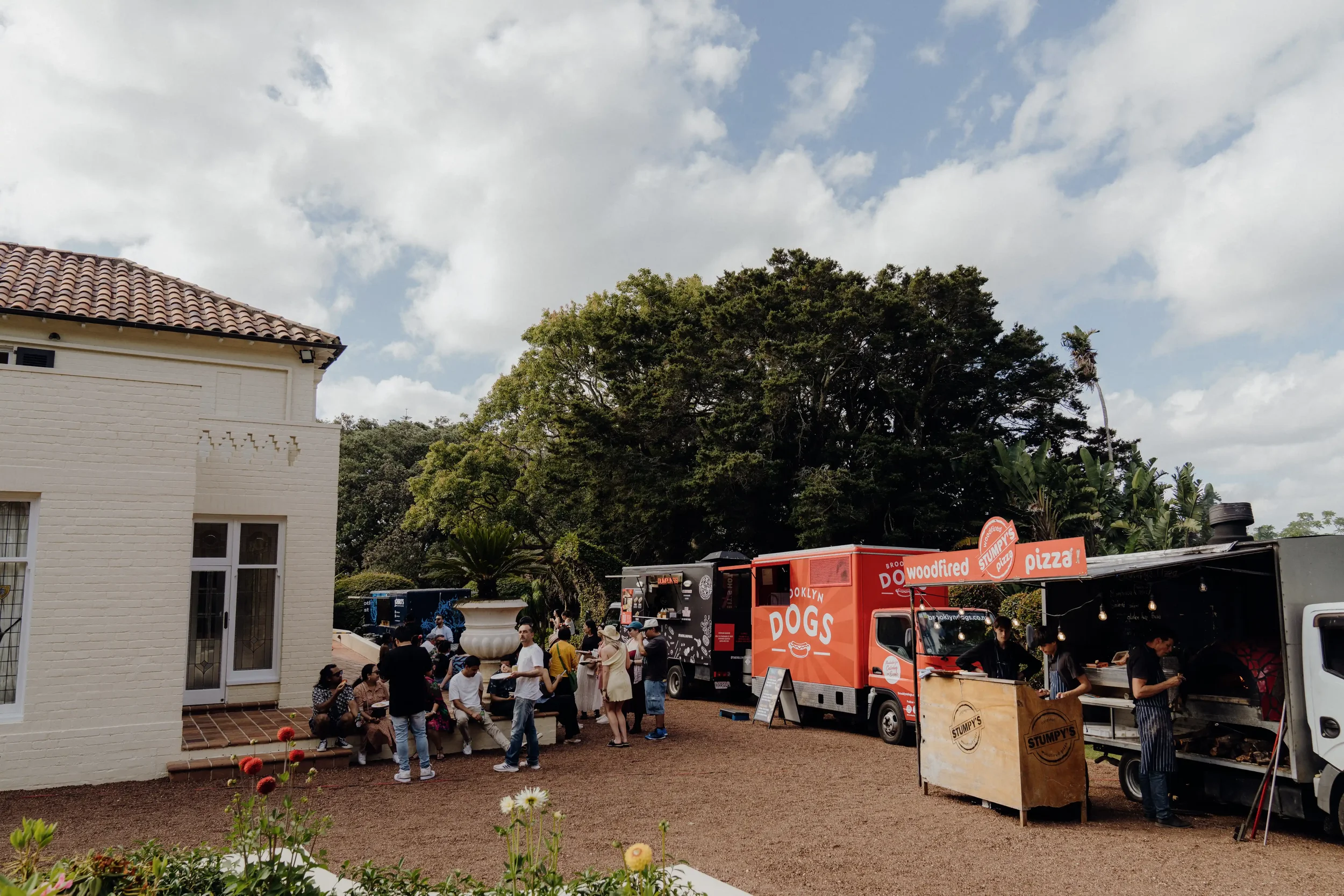 People gathering at food trucks, including ones selling pizza and hot dogs, with a building on the left, trees, and a cloudy sky in the background.