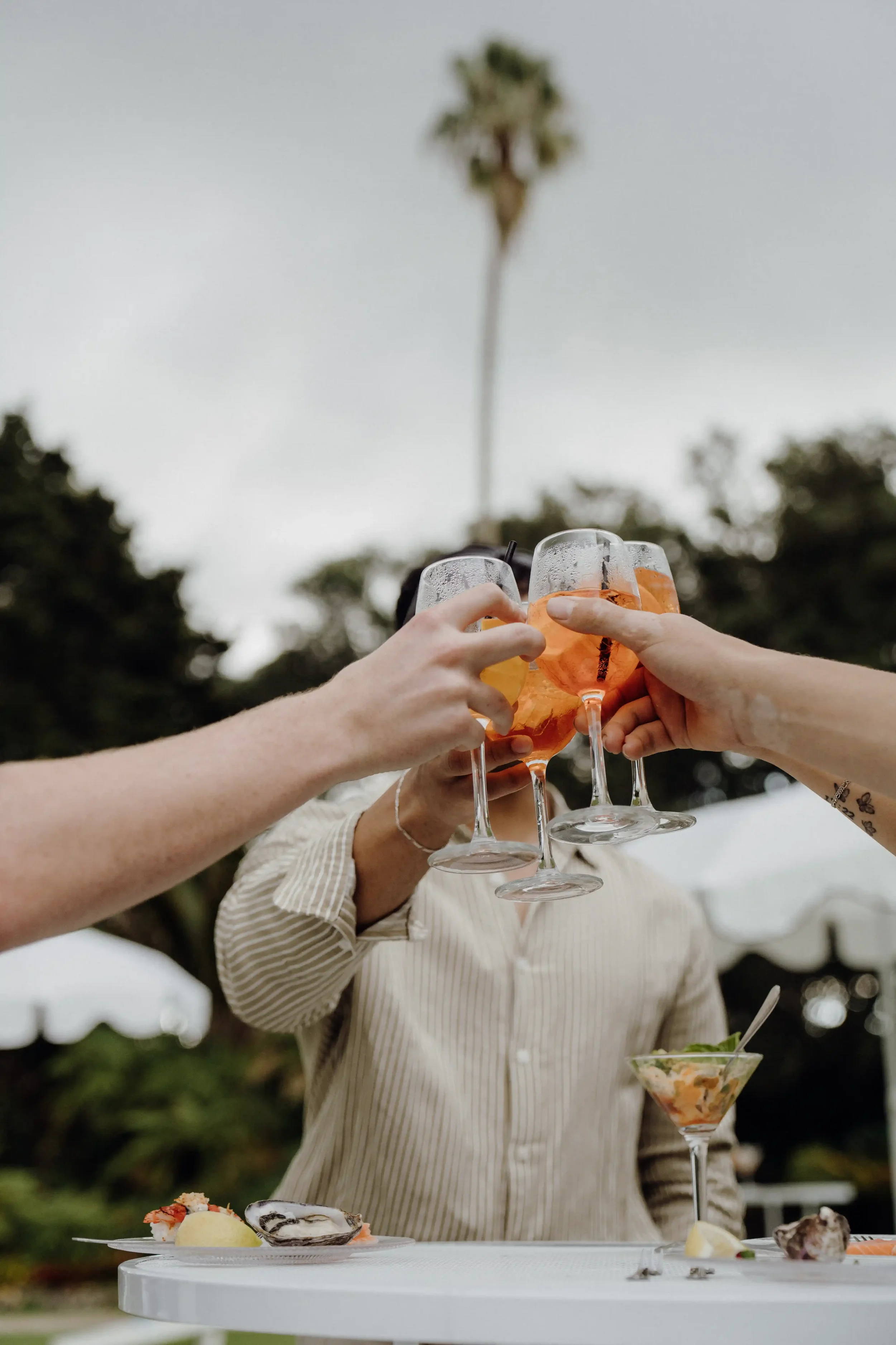 People holding up glasses of orange-colored drinks in a toast, with a blurred outdoor background and plates of food on a white table.