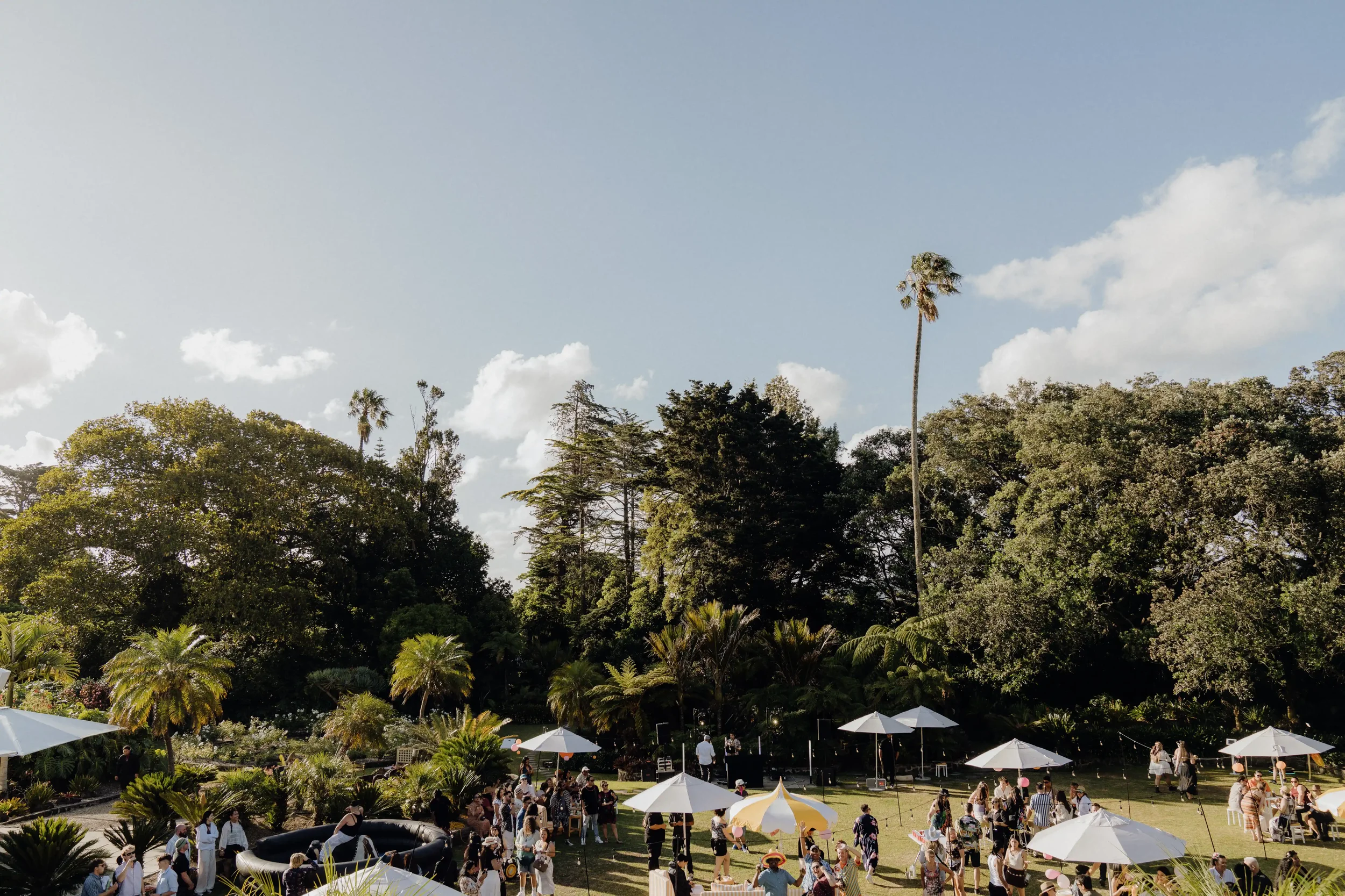 People gathered outdoors in a park or garden with white umbrellas, surrounded by lush trees and greenery under a partly cloudy sky.
