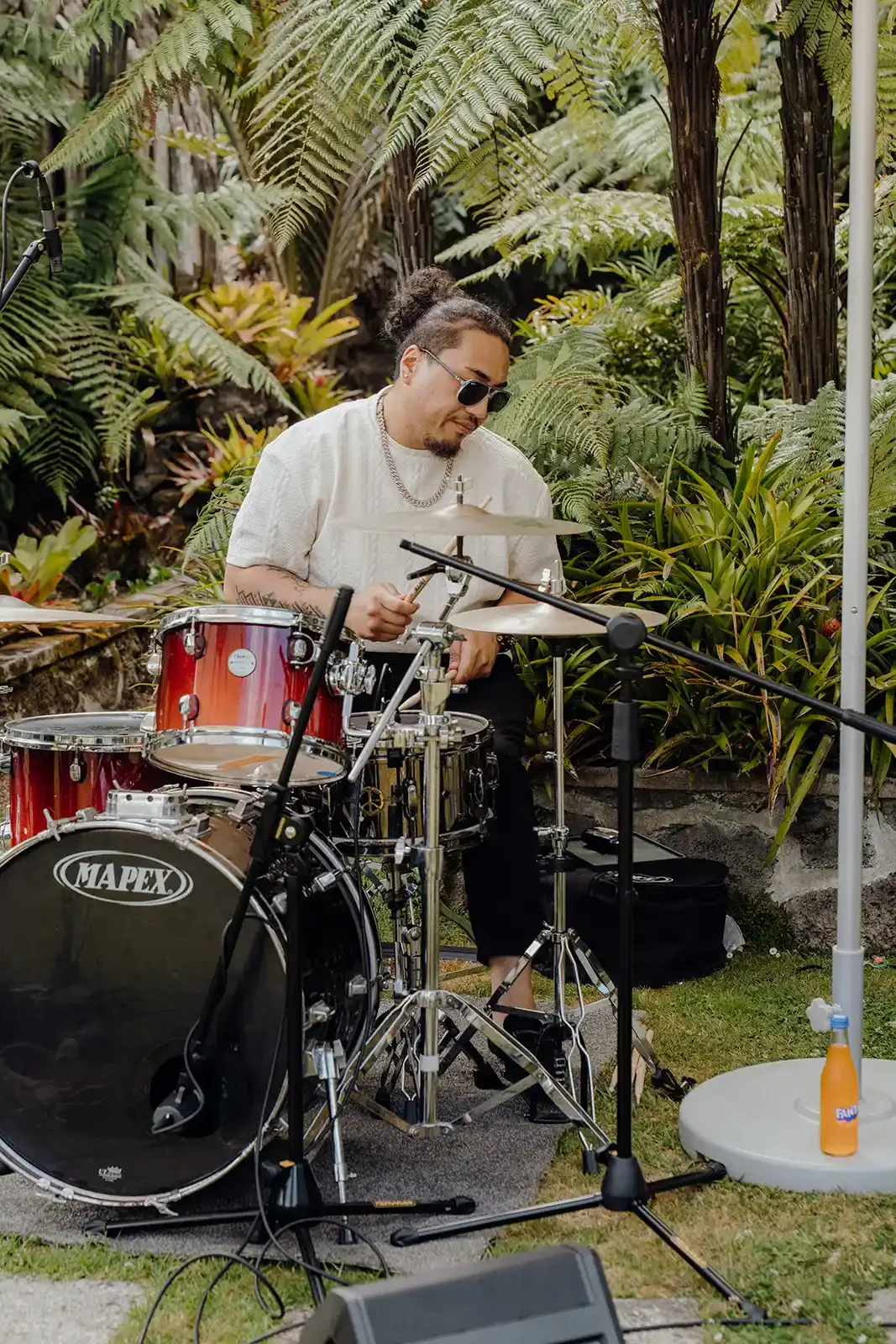 A man with dark hair and sunglasses playing red drums outdoors among lush green tropical plants.