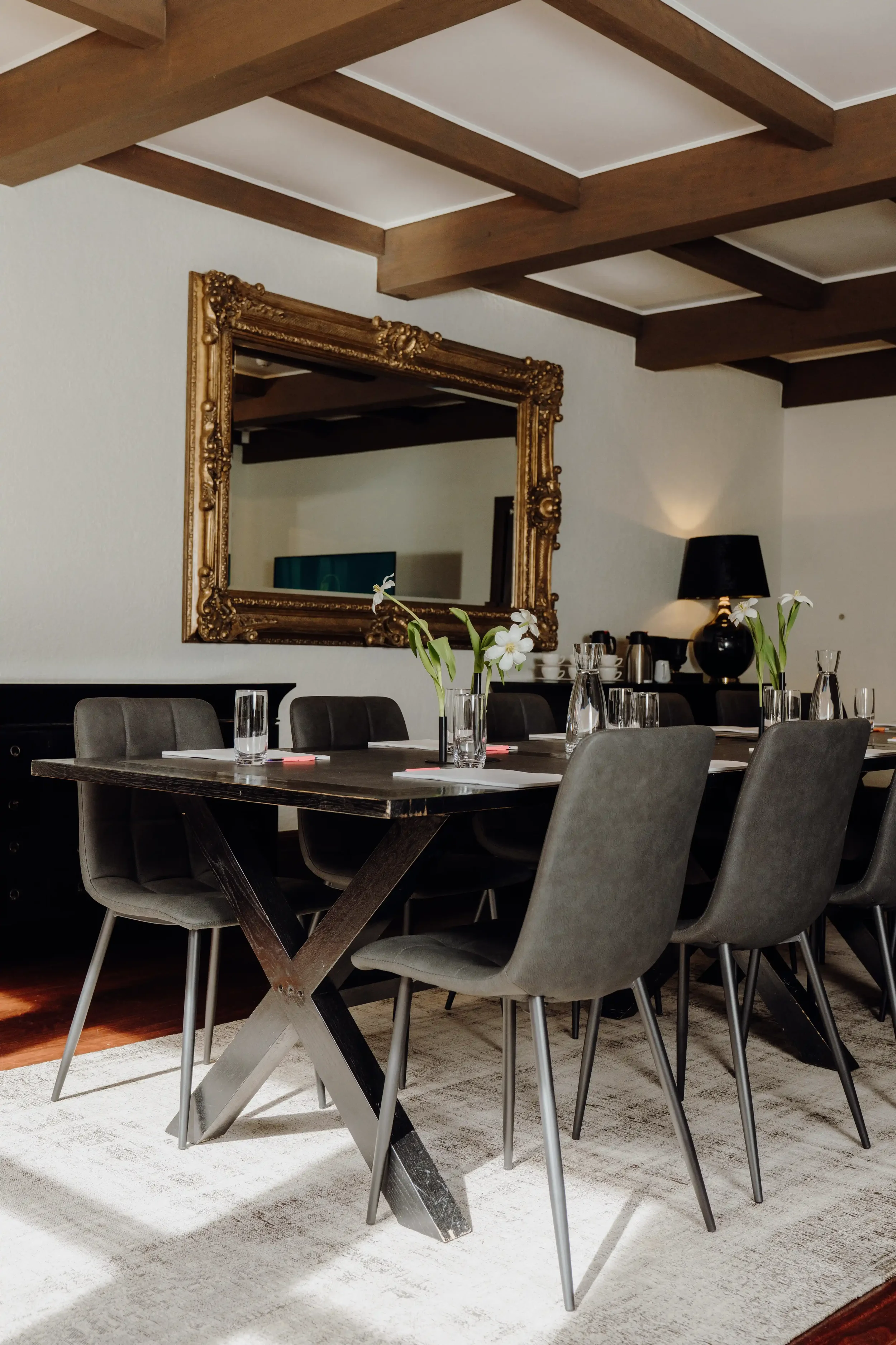 A modern dining room with a dark wooden table and matching gray upholstered chairs. On the table are clear glass vases with white flowers. The room features a large ornate gold-framed mirror on the wall, a black lamp with a shade, and a coffee and te