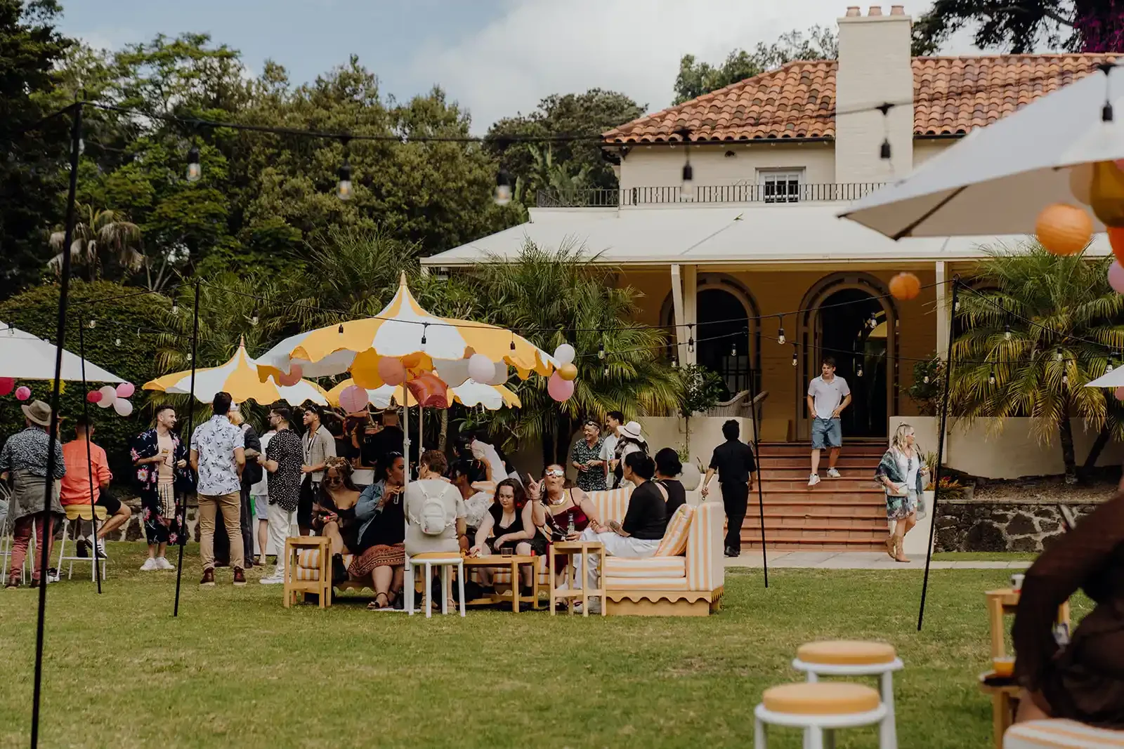 Outdoor party with people socializing, sitting on striped outdoor furniture, under yellow and white umbrellas, decorated with string lights and paper lanterns, in front of a large house with a tiled roof and lush greenery.