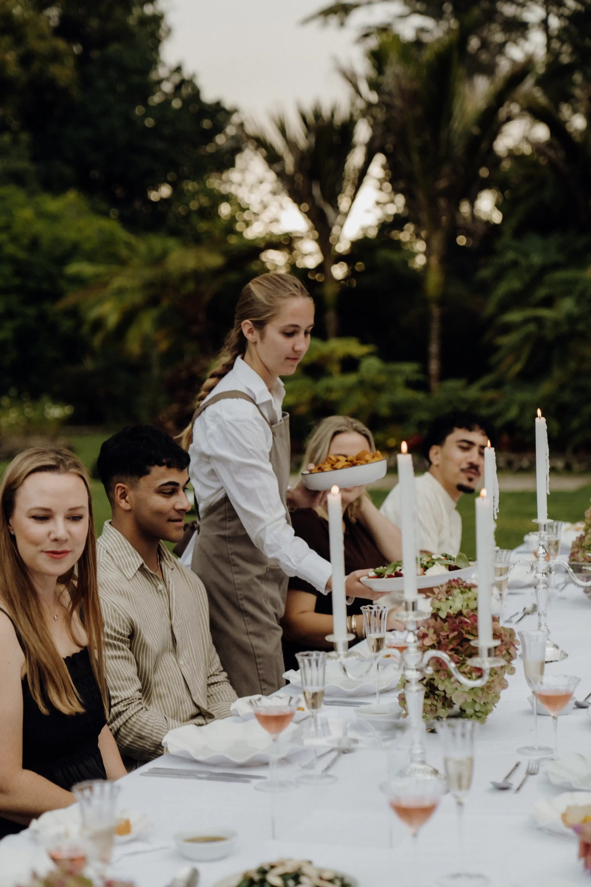 A group of young adults dining outdoors at a long, elegant table decorated with candles, flowers, and glassware, with a woman serving food from a bowl during what appears to be a celebration or event in a lush, green garden.