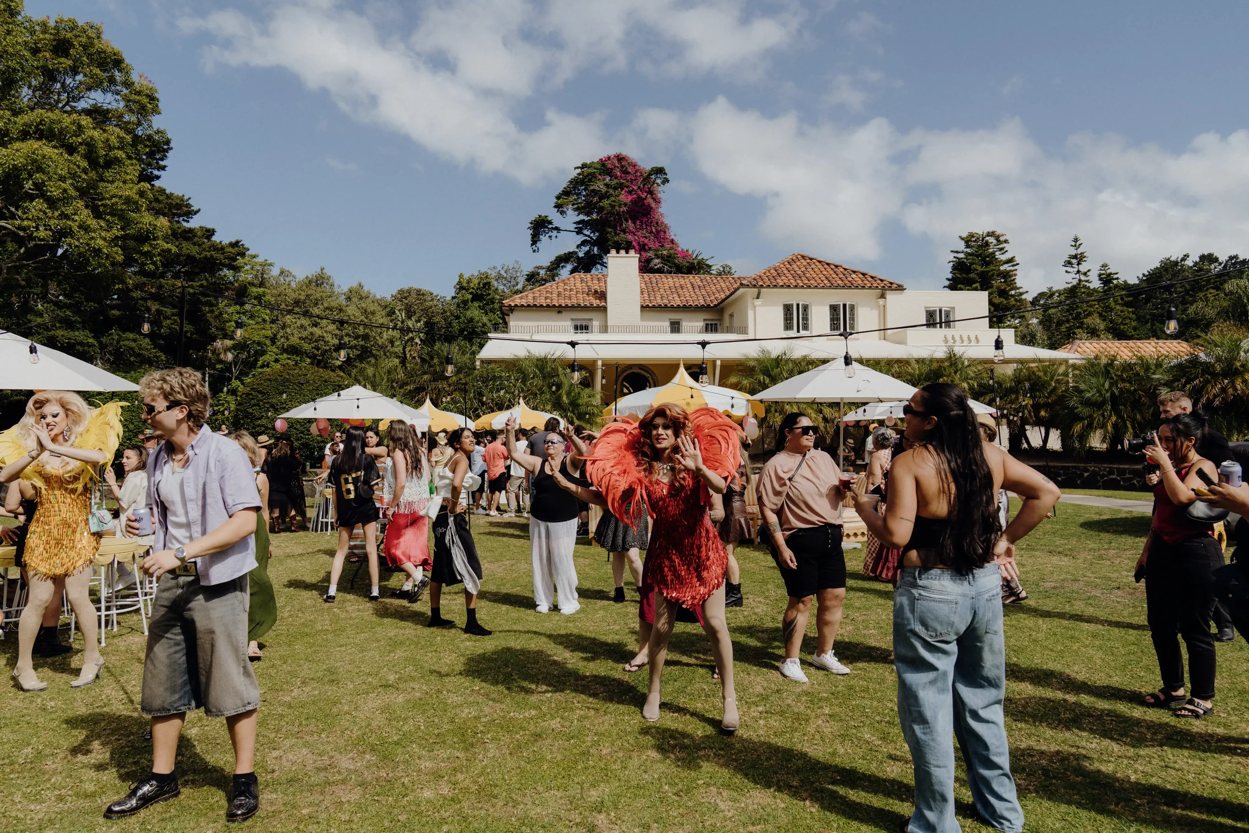 People gathered at an outdoor event on a sunny day, with some dressed in colorful costumes, and white umbrellas providing shade. A large house with a red-tiled roof and a chimney is visible in the background, surrounded by trees and a clear blue sky 