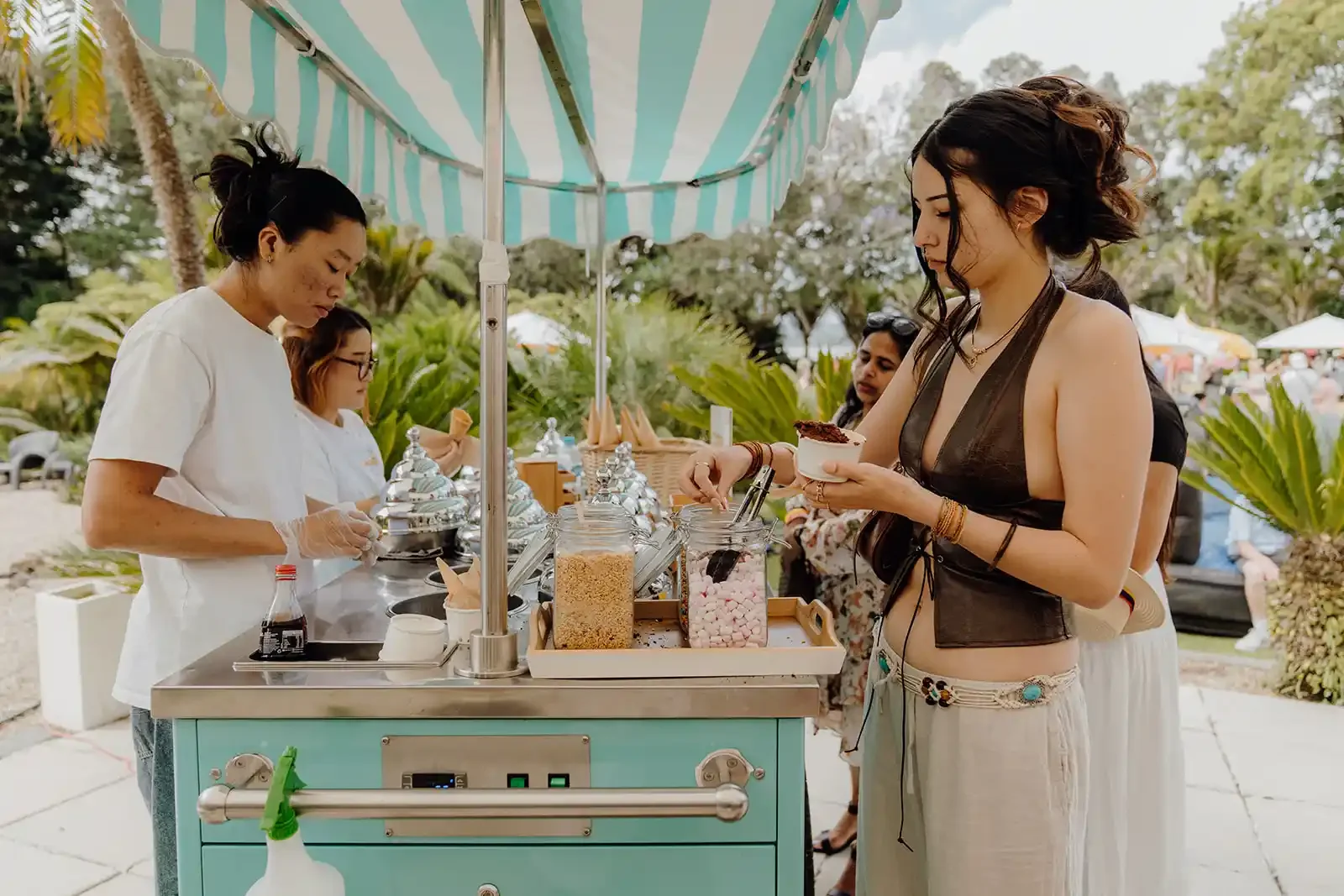 A woman in a black top and beige skirt is serving herself ice cream from a vendor cart, with a line of people behind her.