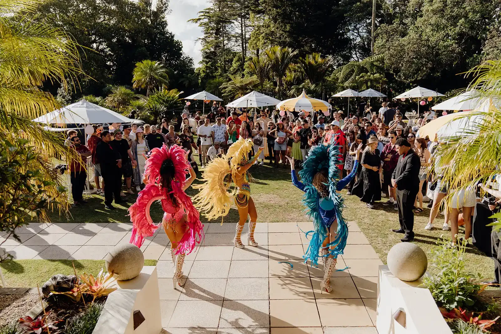 Three dancers in colorful feather costumes perform for a crowd at an outdoor celebration. The audience watches under umbrellas amidst trees and greenery.