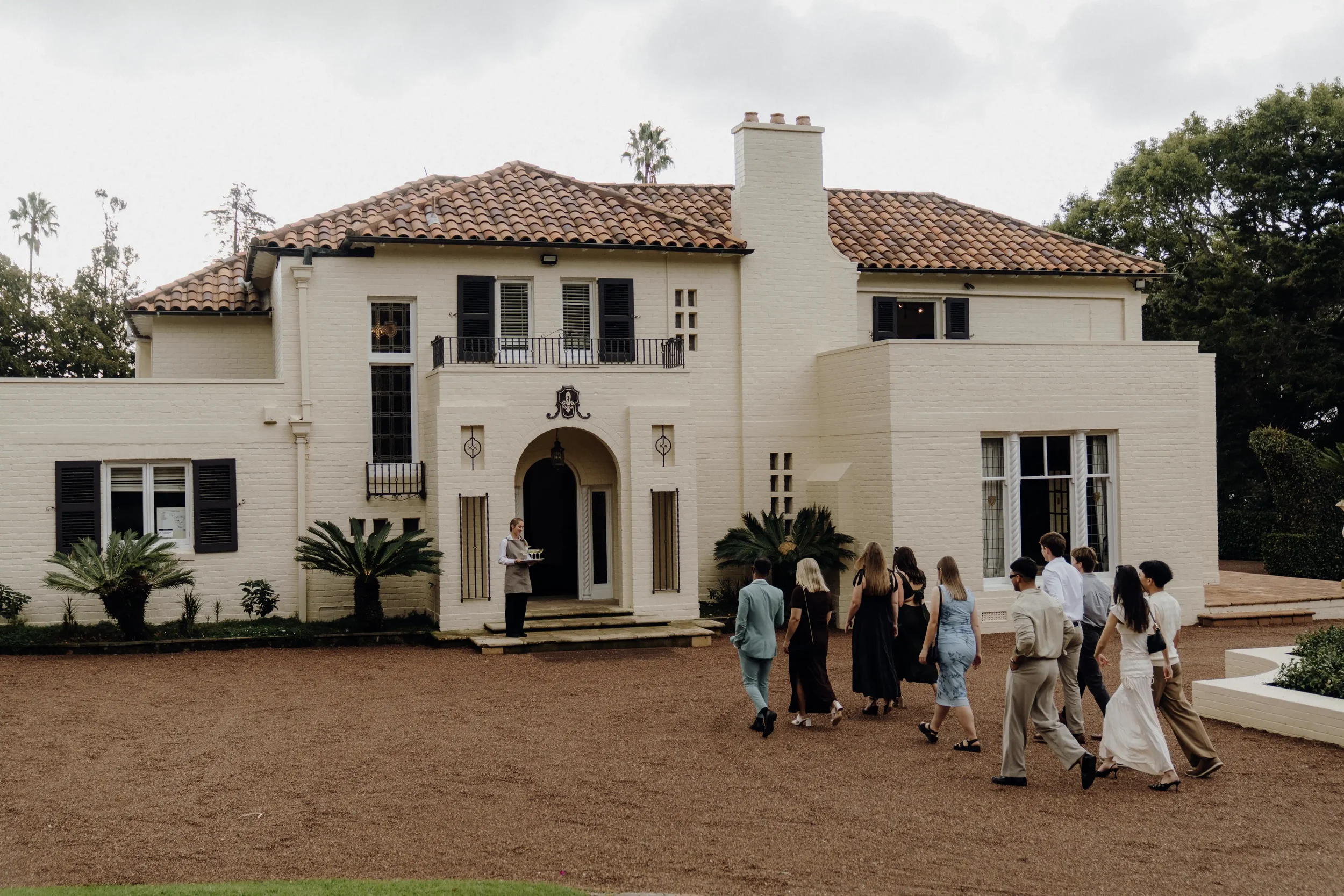 A group of people walking towards a large, white, two-story house with a tiled roof, black shutters, and a front arched door, with a woman standing at the entrance holding a tray, and trees and cloudy sky in the background.